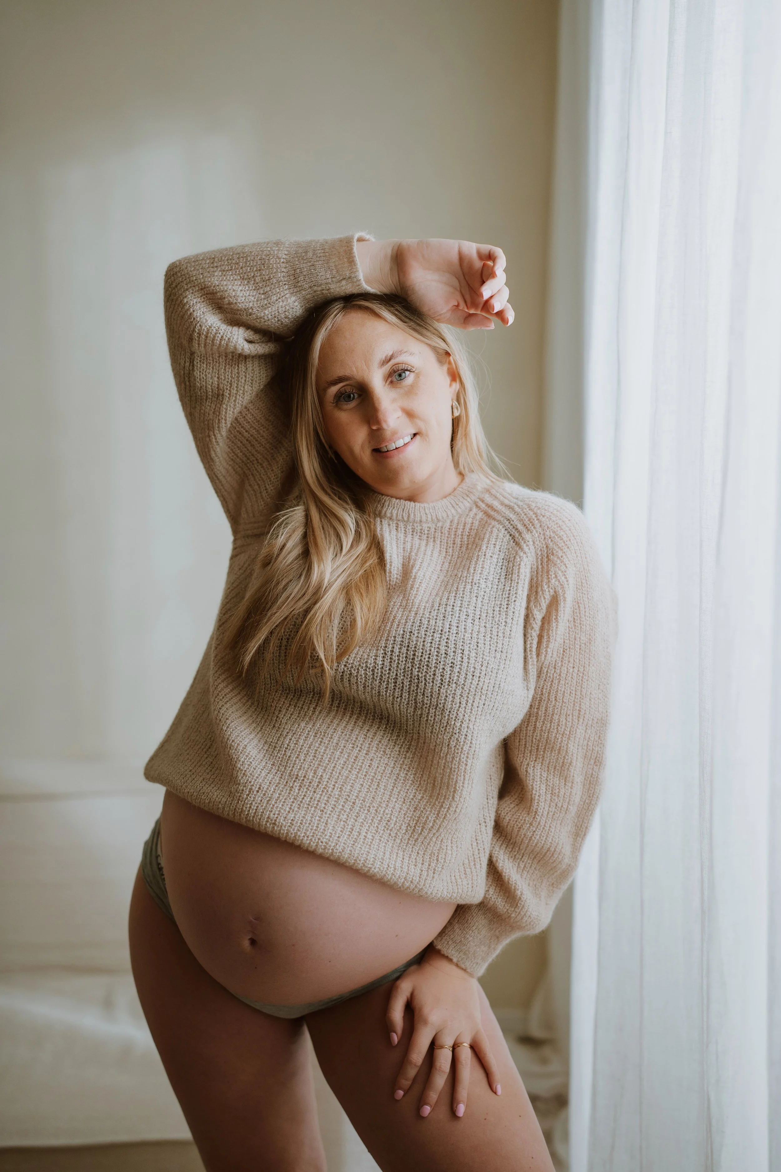 A pregnant woman with long blonde hair, wearing a beige sweater and black underwear, standing indoors near a window with sheer curtains, holding her thigh and looking at the camera.