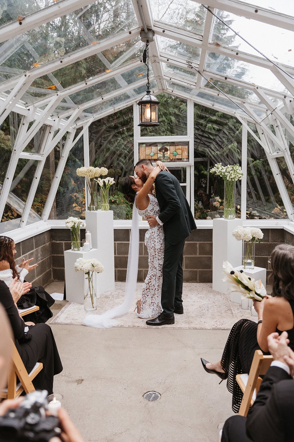 A bride and groom kissing during their wedding ceremony in a glass greenhouse with floral arrangements and guests watching.