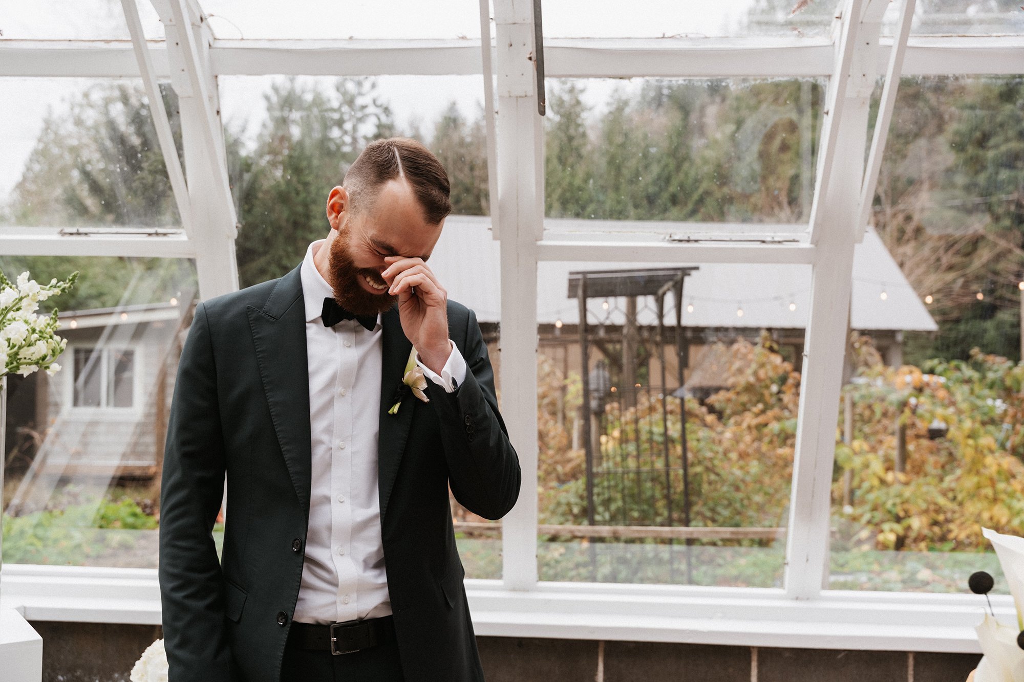 A man in a tuxedo with a bow tie is laughing and covering his face with his hand, standing in a greenhouse or glass structure with an outdoor garden visible behind him.