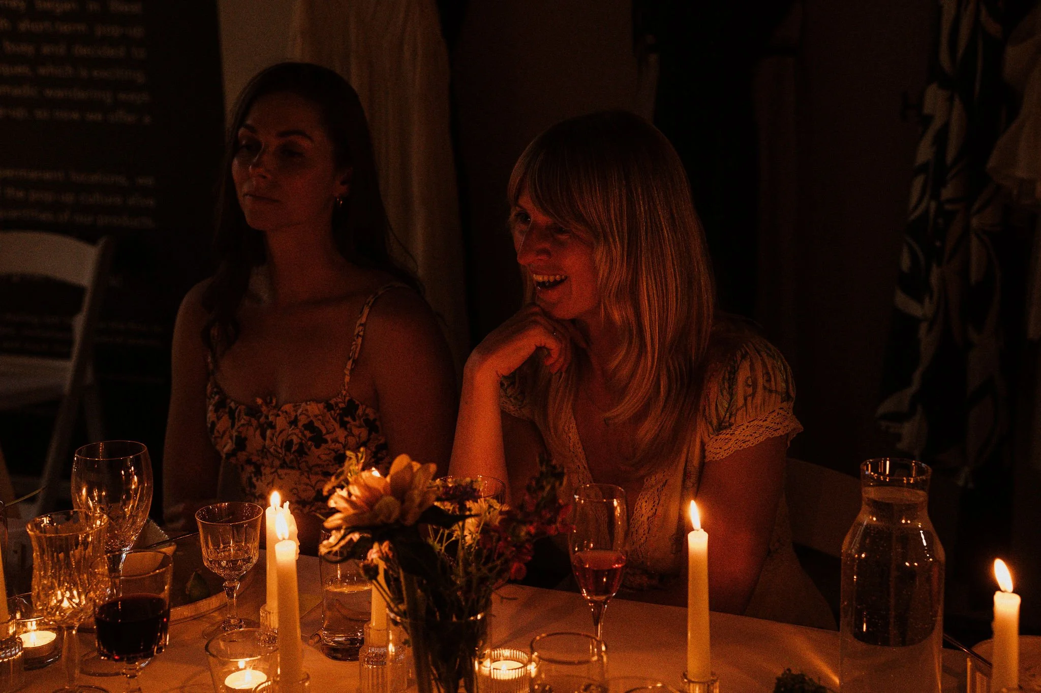 Two women sitting at a dinner table lit by candles, with one woman smiling and the other looking away, in a dimly lit setting.