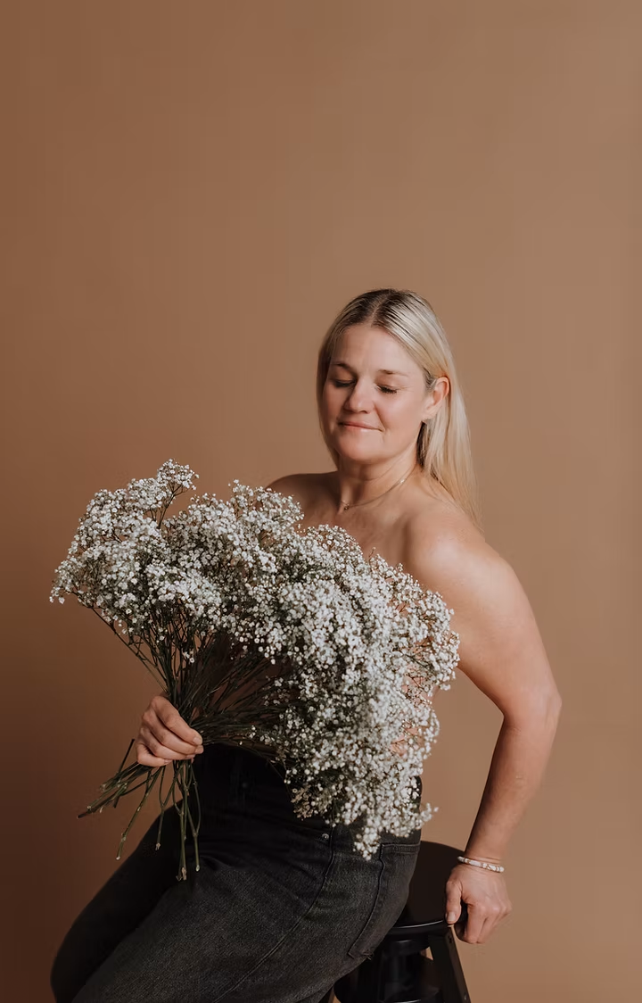 A woman with blonde hair seated on a stool, holding a large bouquet of white baby's breath flowers against a plain brown background, wearing black jeans.