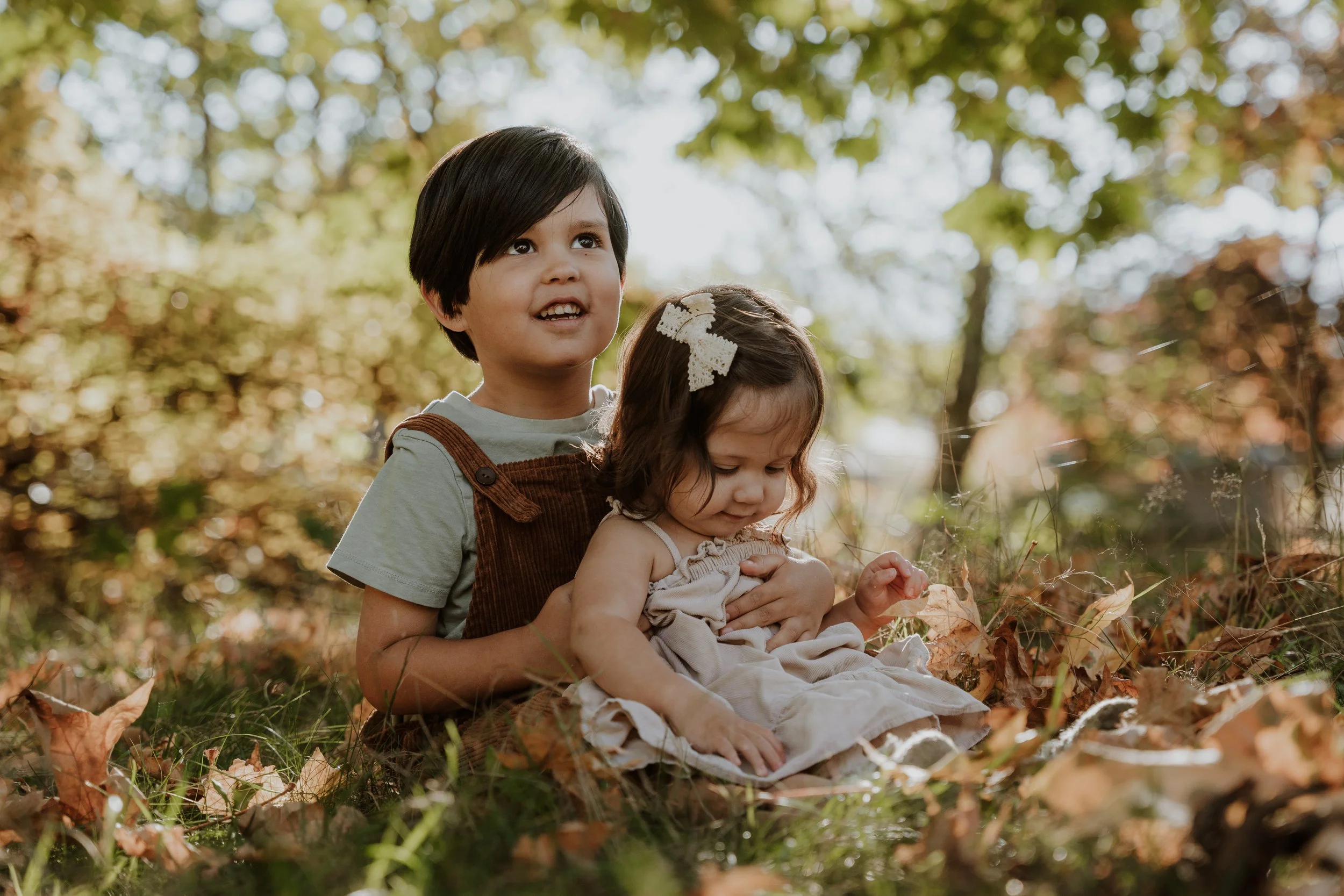 Two children, a boy and a girl, sitting outdoors among fallen leaves in a forest during autumn, with the boy holding the girl from behind.
