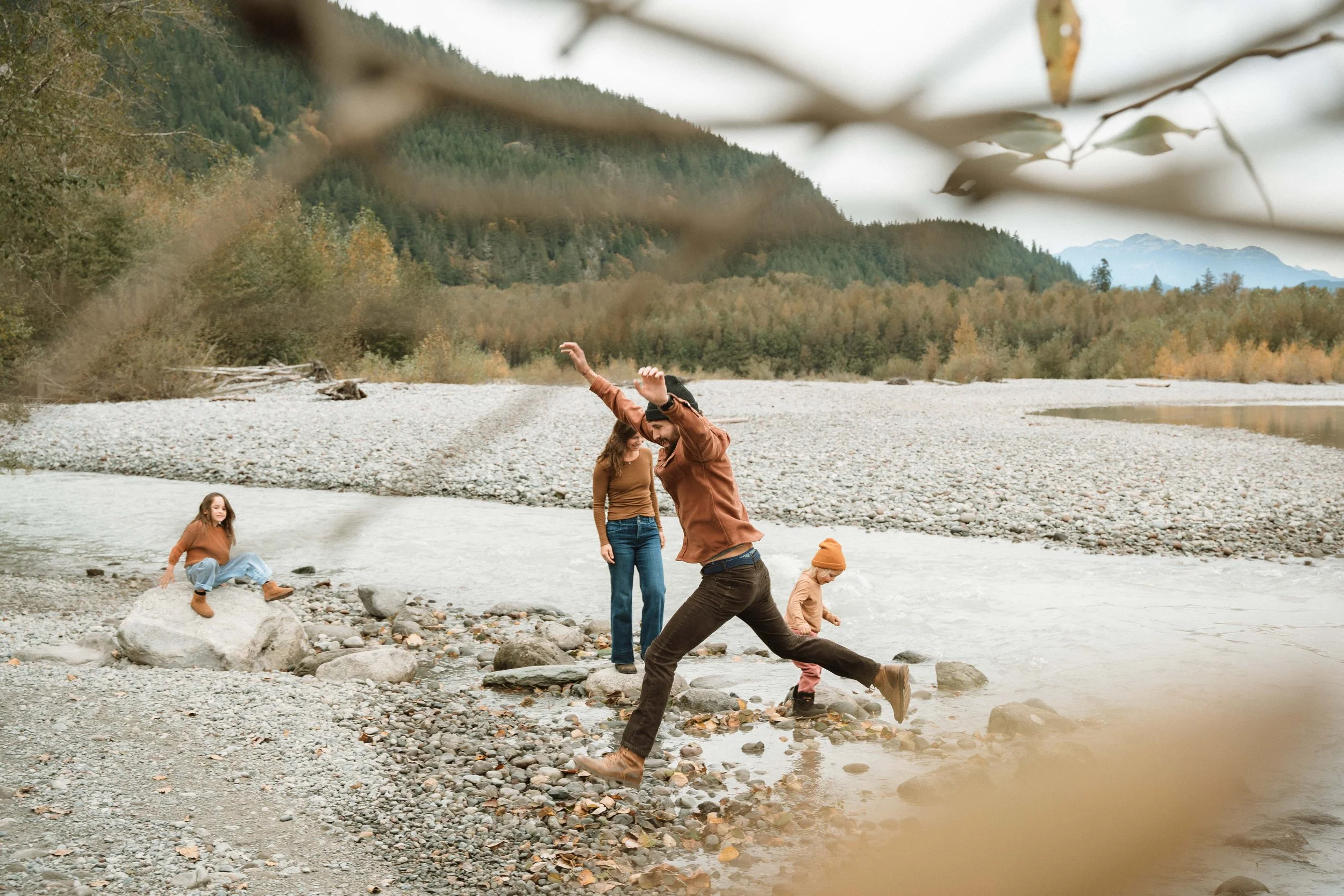 Family outdoors by river in mountains, a woman and two children playing and jumping on rocks, with a girl sitting on a large rock nearby.