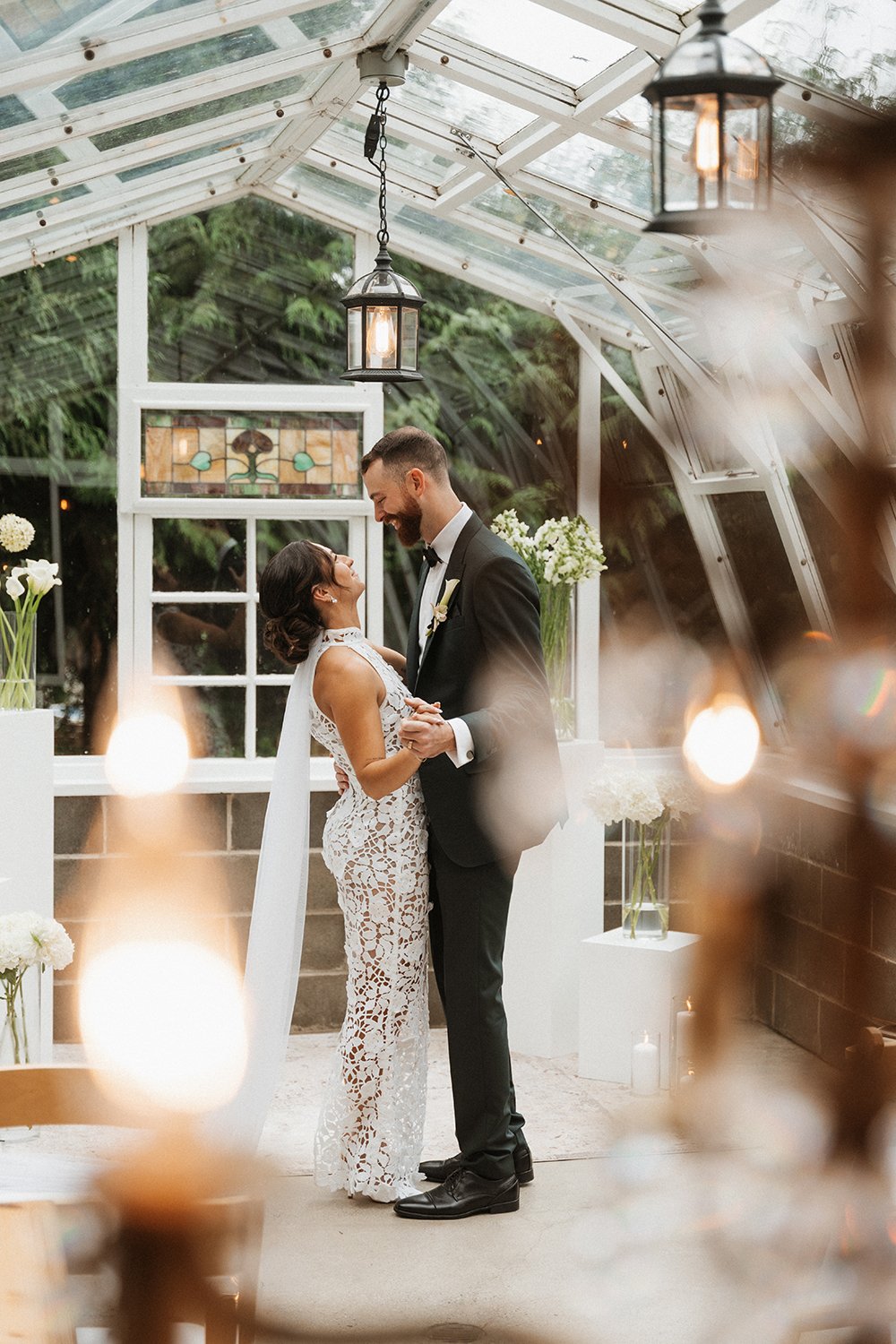 A bride and groom dancing inside a glass greenhouse, surrounded by vases of white flowers and warm hanging lights.