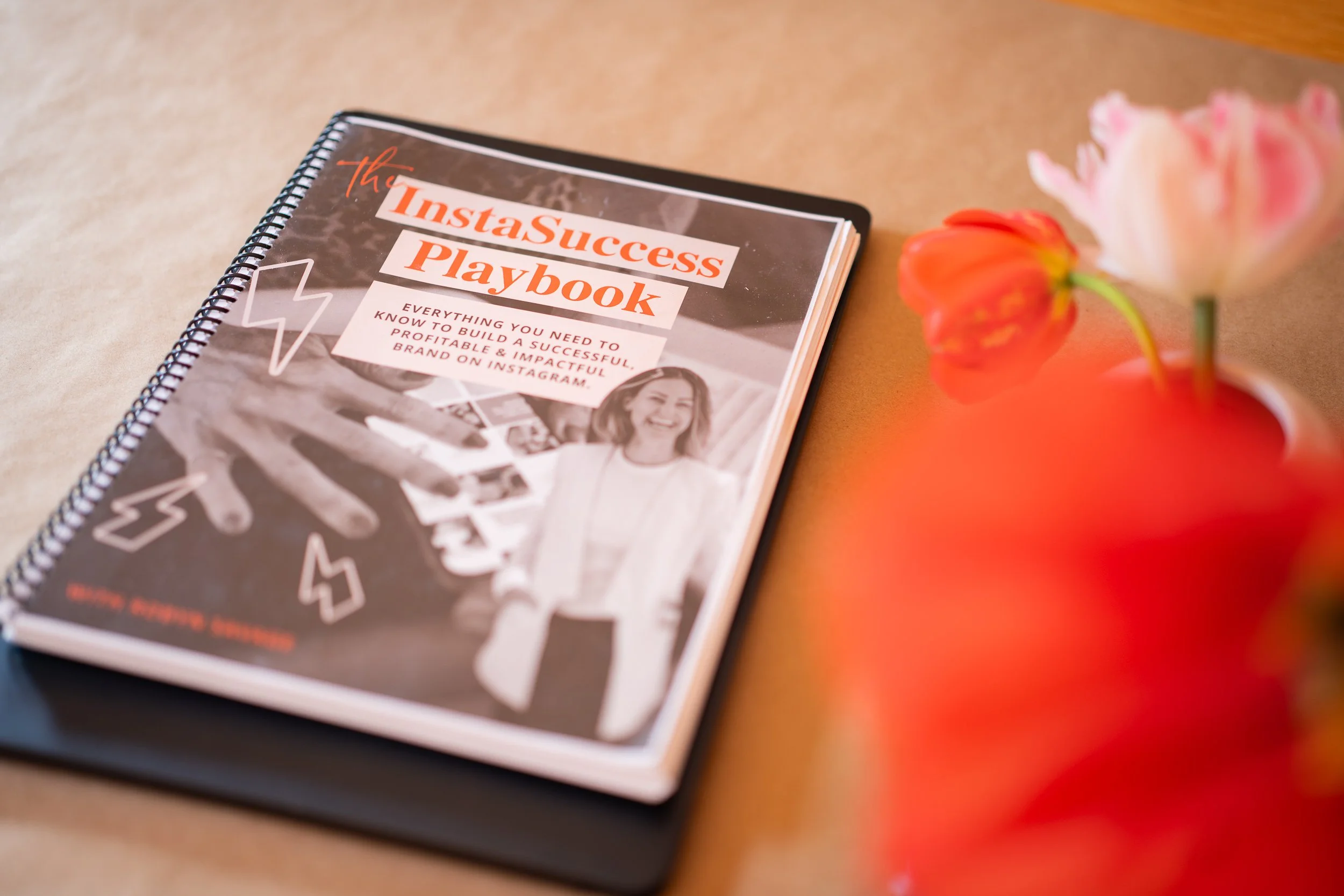 A spiral notebook titled 'InstaSuccess Playbook' resting on a black folder, with pink and red tulips in a vase nearby, on a wooden surface.