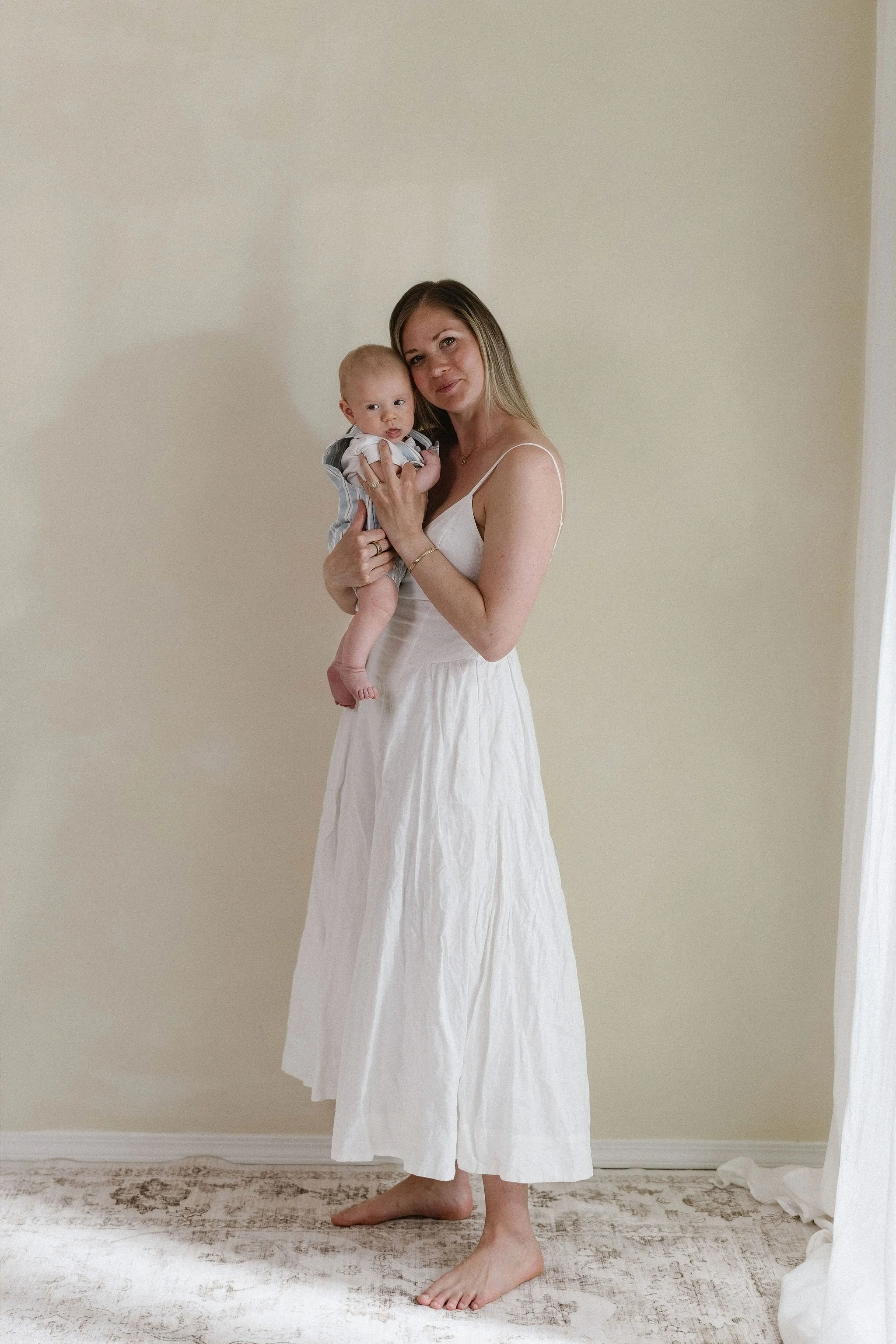 A woman standing barefoot on a patterned rug, holding a baby, wearing a white dress with thin straps, in a room with beige walls and a white curtain.
