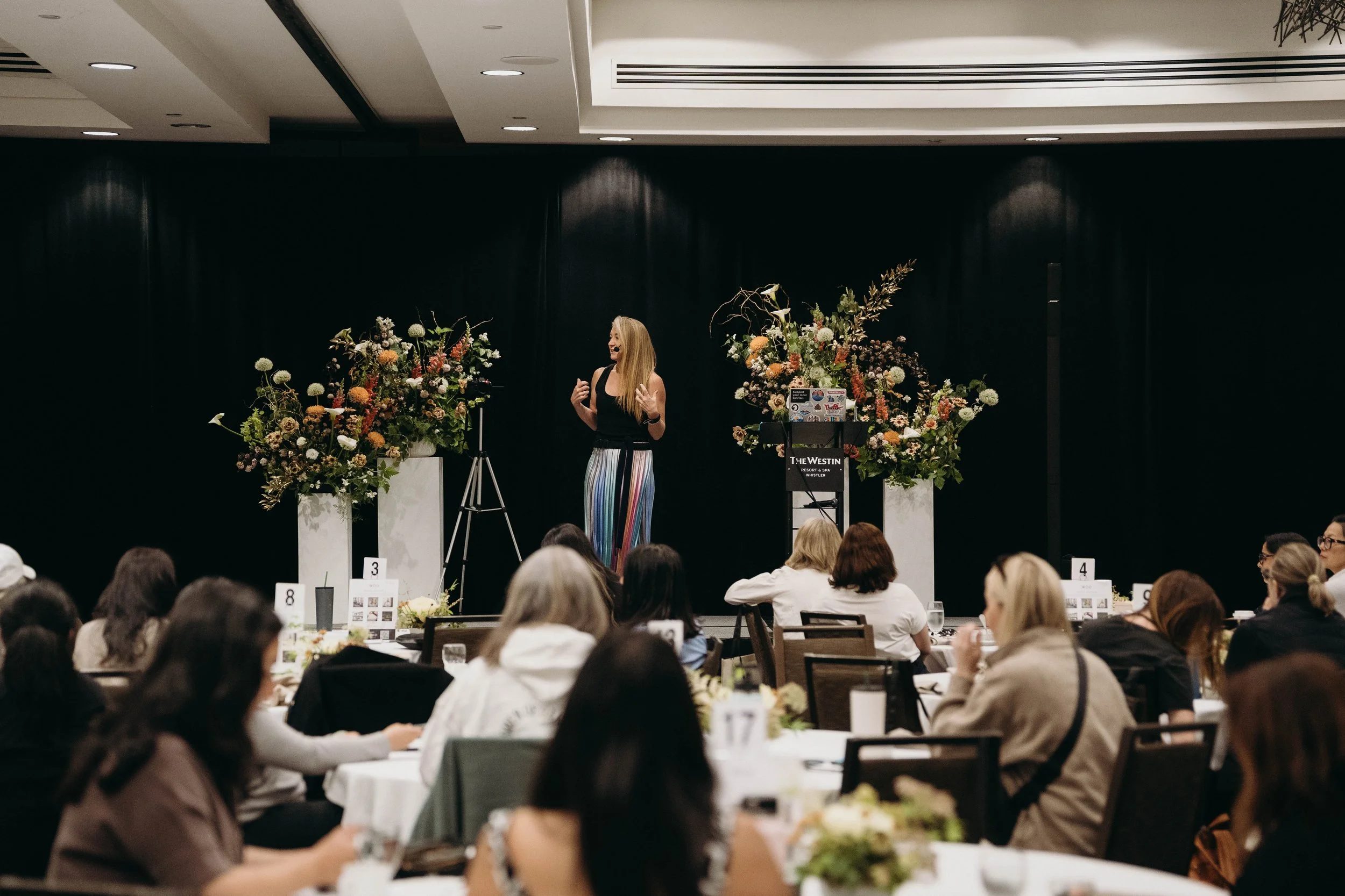 A woman in a black top and colorful striped pants giving a presentation on stage at a conference with floral arrangements on either side. The audience is seated at round tables in a hotel event space.