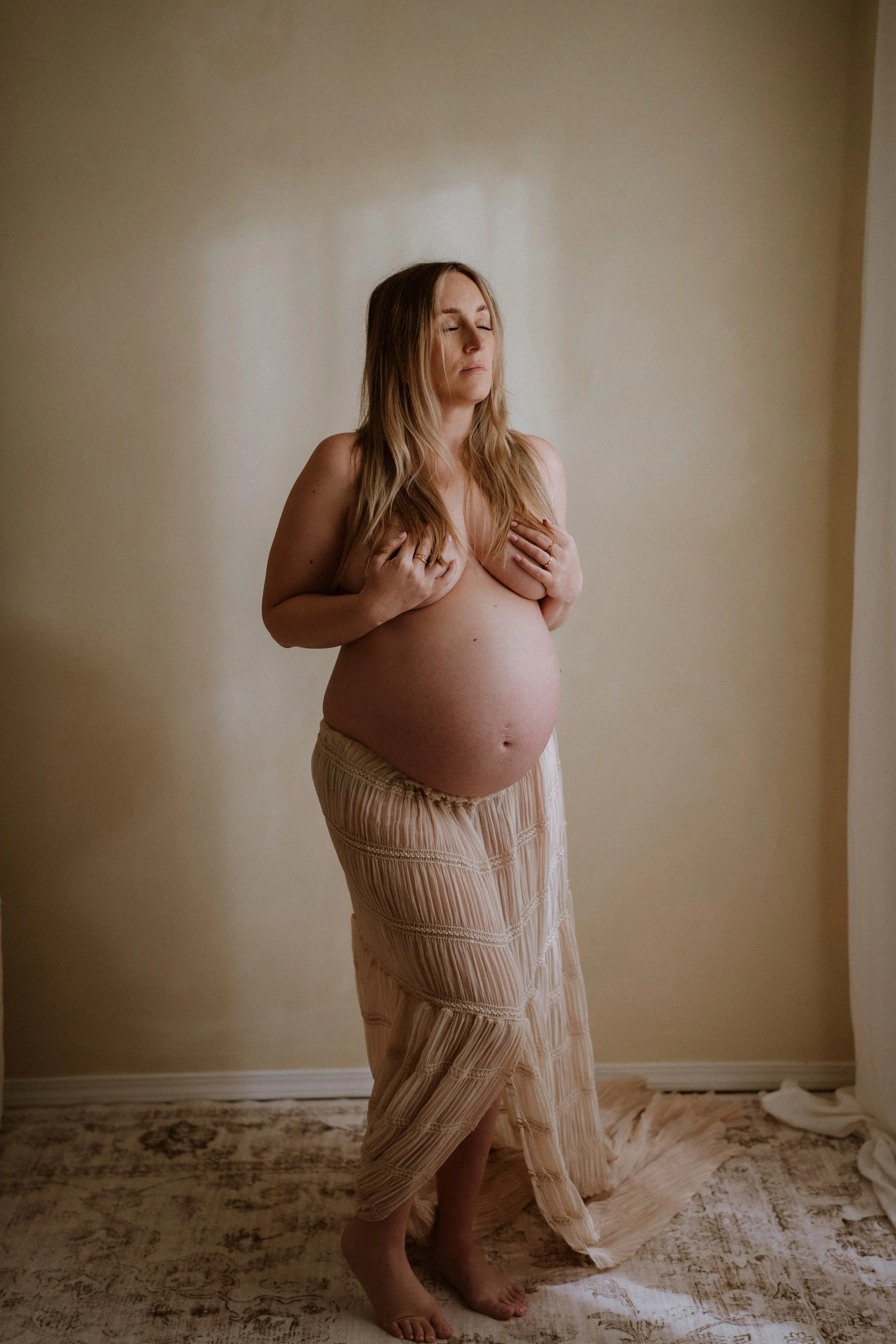 A pregnant woman standing barefoot on a patterned rug in front of a plain beige wall, with her eyes closed and hands resting gently on her breast, wearing beige pants and a light-colored skirt.