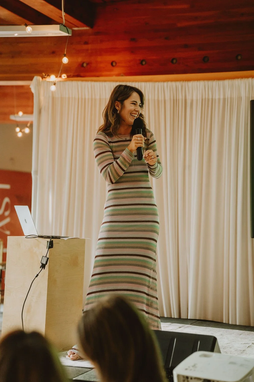 A woman in a long, striped dress and earrings is holding a microphone and smiling on stage, with a beige curtain backdrop and a small table with a laptop nearby.
