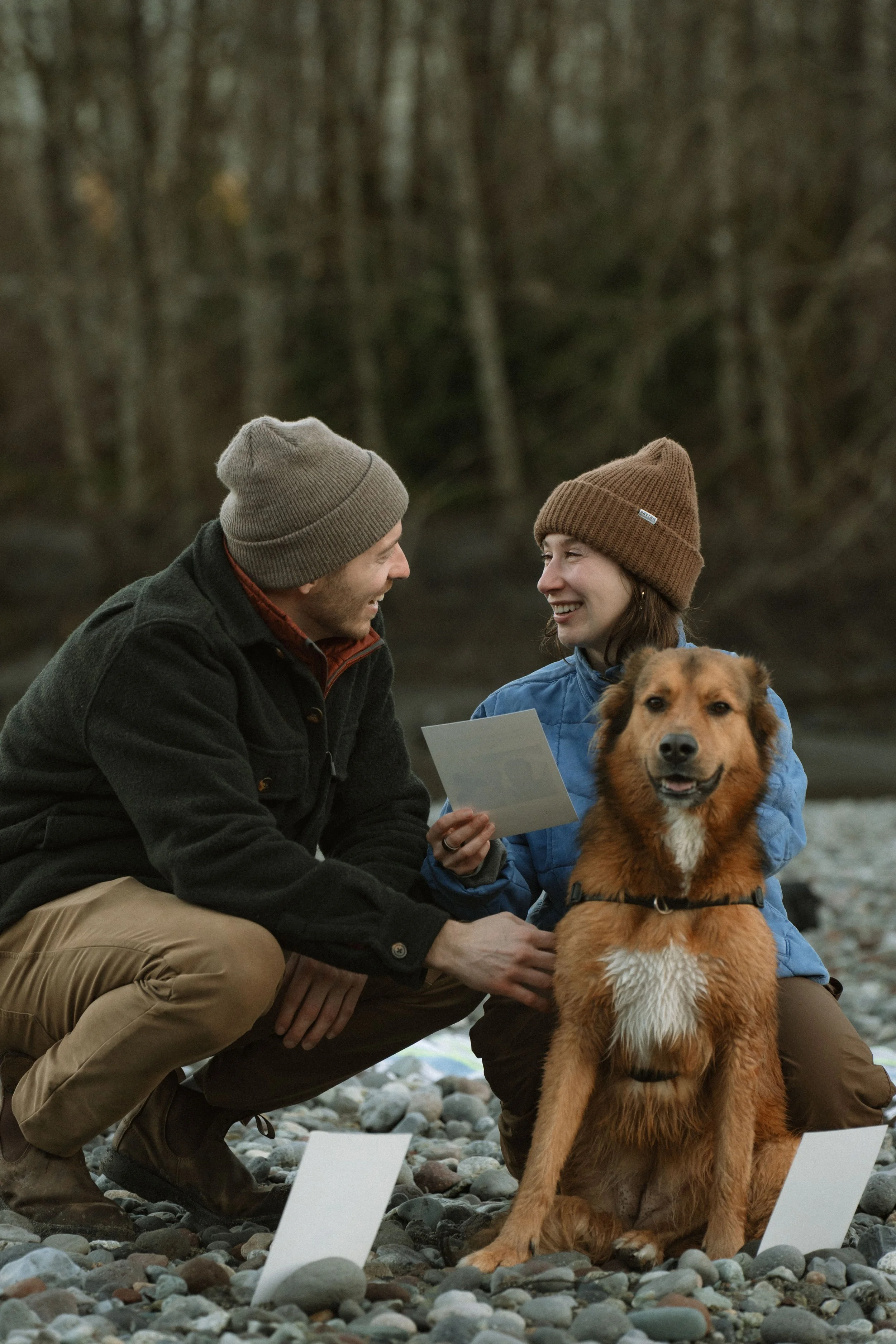 Two people wearing beanies and outdoor jackets are kneeling on a rocky riverbank, smiling and looking at each other, with a brown dog sitting in front of them. The woman is holding a card or photograph. There is trees and a blurred outdoor background