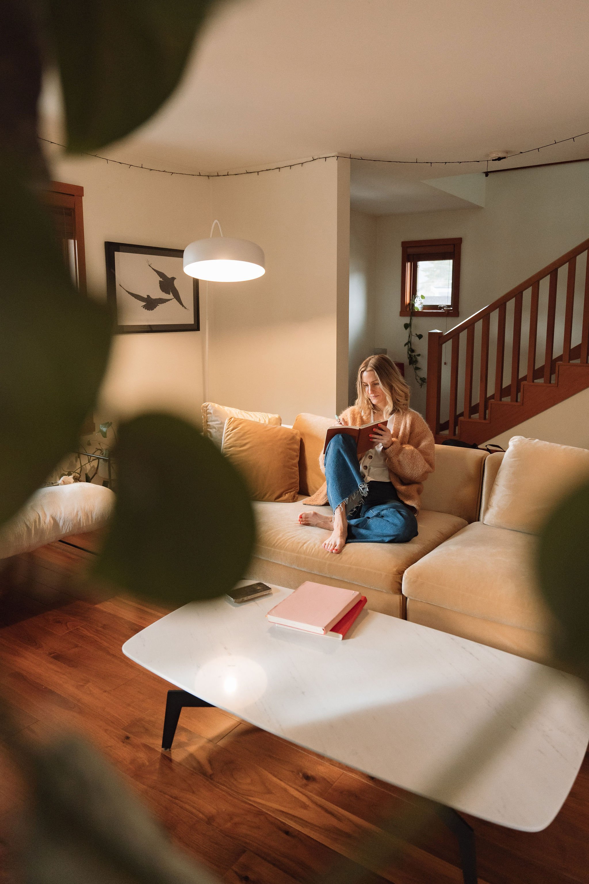A cozy living room with a woman sitting on a beige couch reading a book, surrounded by warm lighting, including a ceiling lamp, and decorated with framed bird artwork on the wall and string lights on the ceiling. There is a small window with a plant 