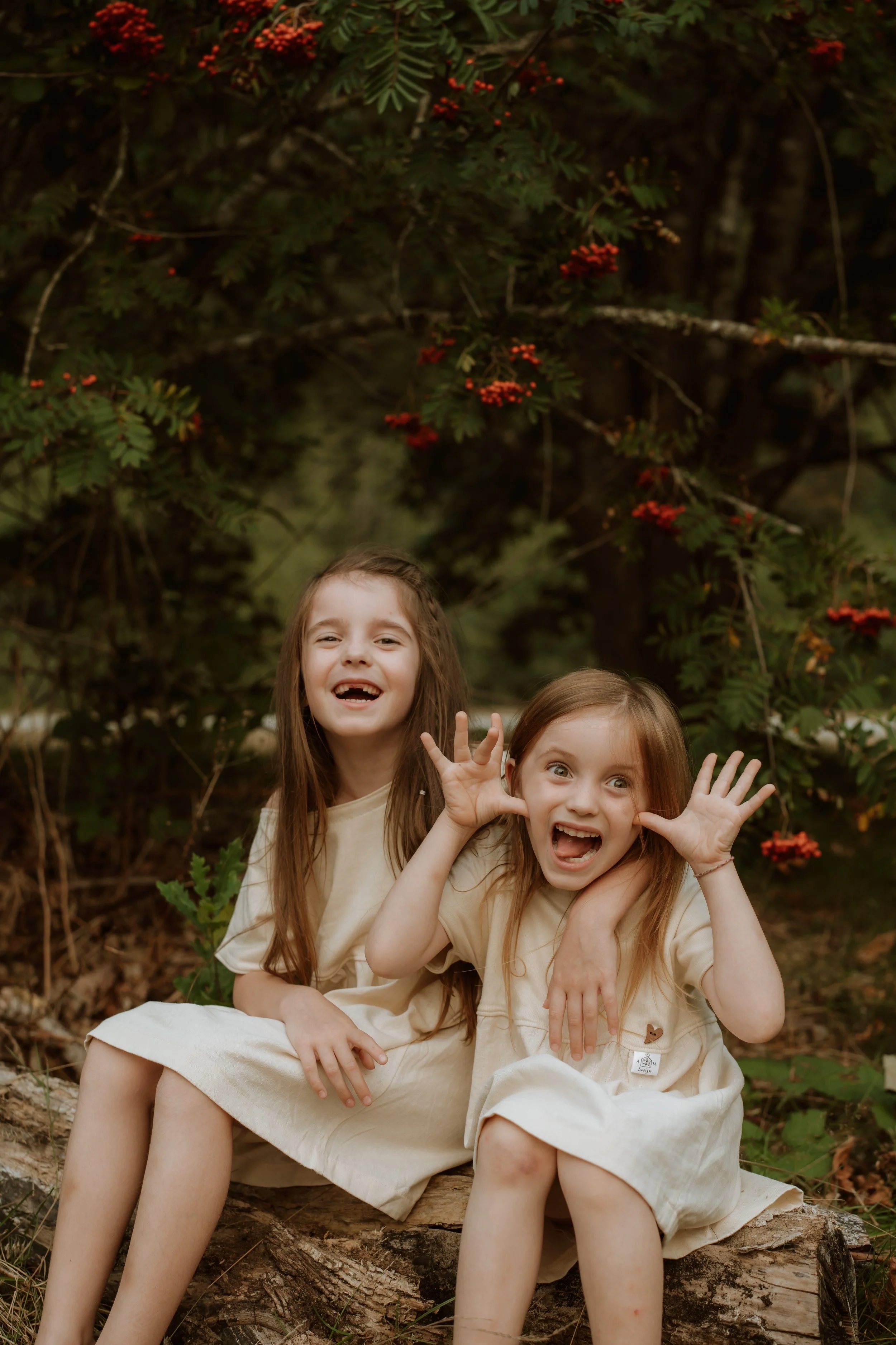 Two young girls sitting on a fallen log in a forest, smiling and making funny faces. One girl has her hand near her face with fingers out, and the other girl is smiling. They are surrounded by dense greenery with red berries on the trees.