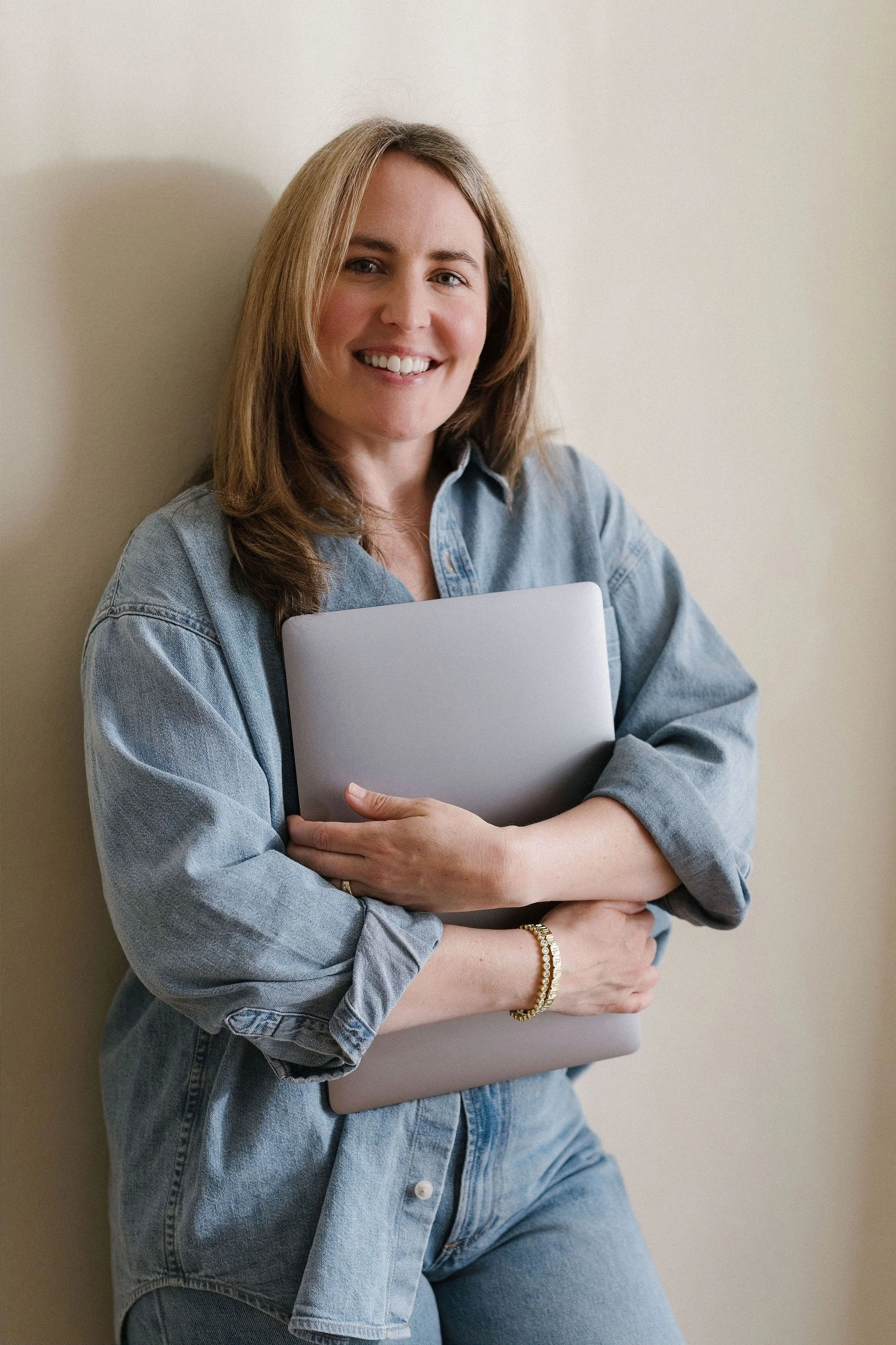 A woman with shoulder-length light brown hair, wearing a light blue denim shirt and jeans, smiling while holding a laptop and standing against a plain beige wall.
