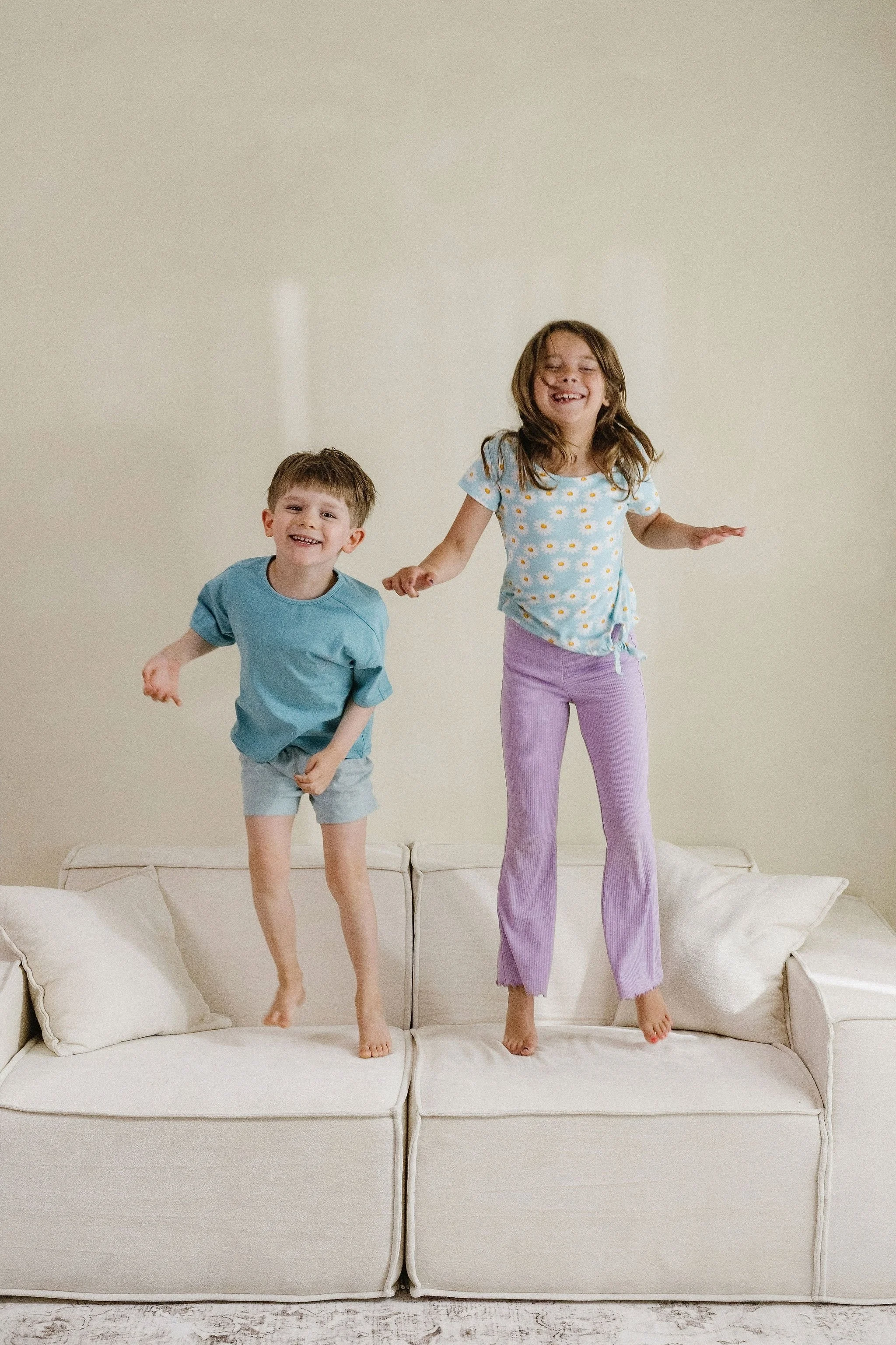 Two children jumping on a white couch, a boy and a girl, smiling and enjoying themselves in a room with plain beige walls.