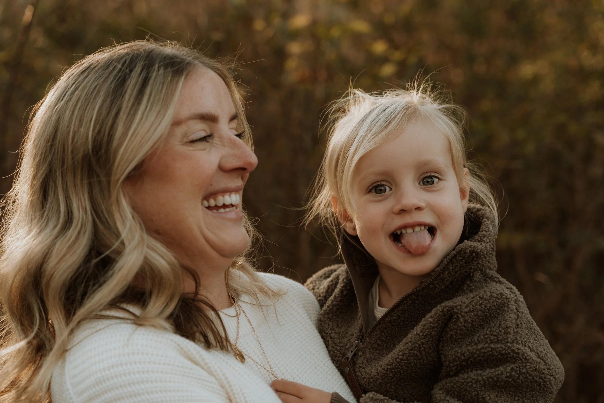 A woman holding a young girl outdoors during autumn, both smiling and expressing happiness.