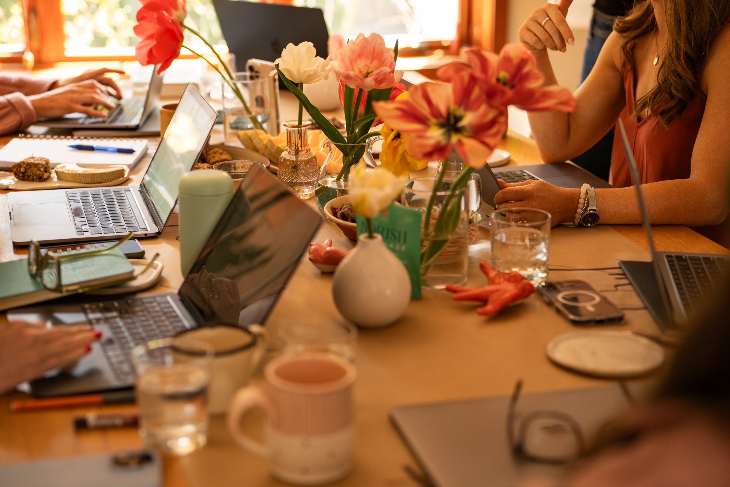 A group of people working on laptops around a wooden table with flowers and snacks, some with glasses of water and a cup of tea.