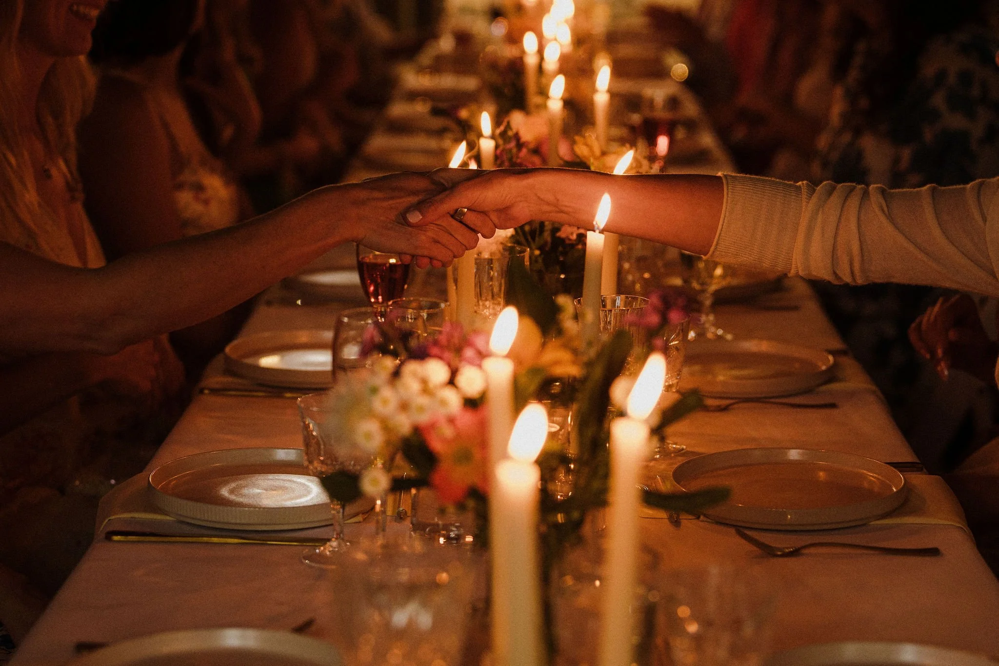A dinner table decorated with candles and flowers, where two people are holding hands across the table in a warm, intimate setting.