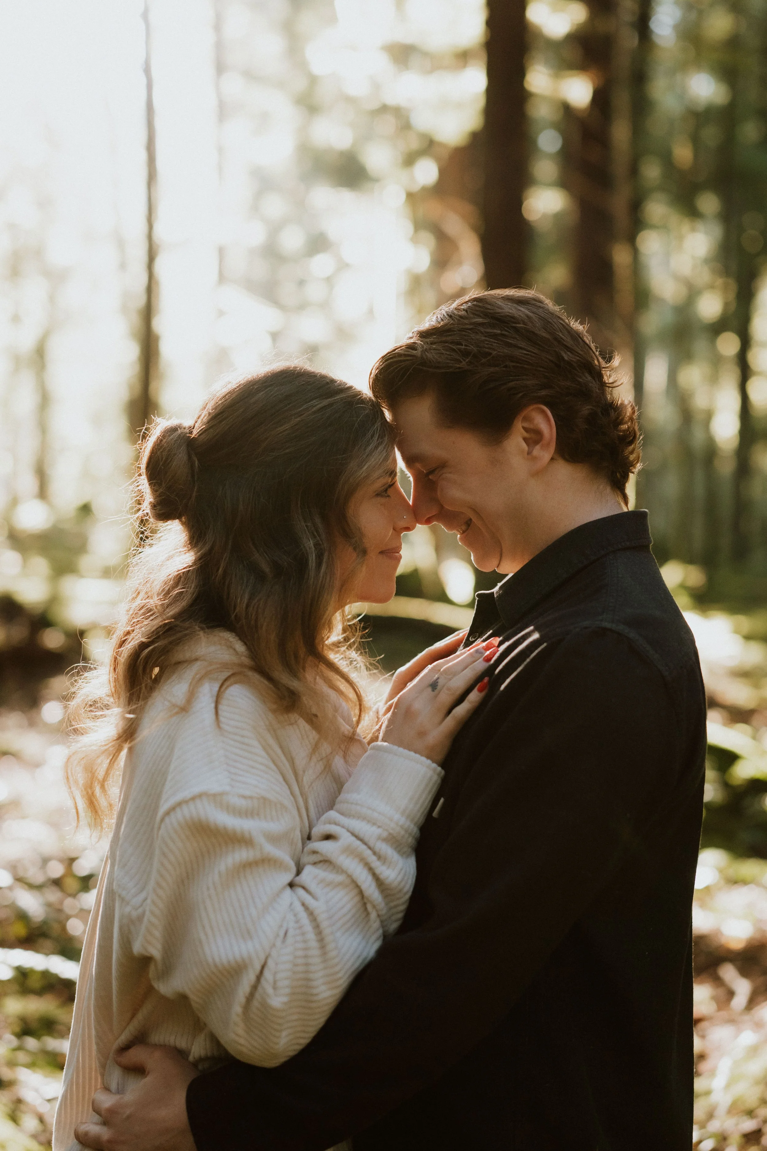 A couple stands close in a forest, touching foreheads and holding each other, with sunlight filtering through trees in the background.