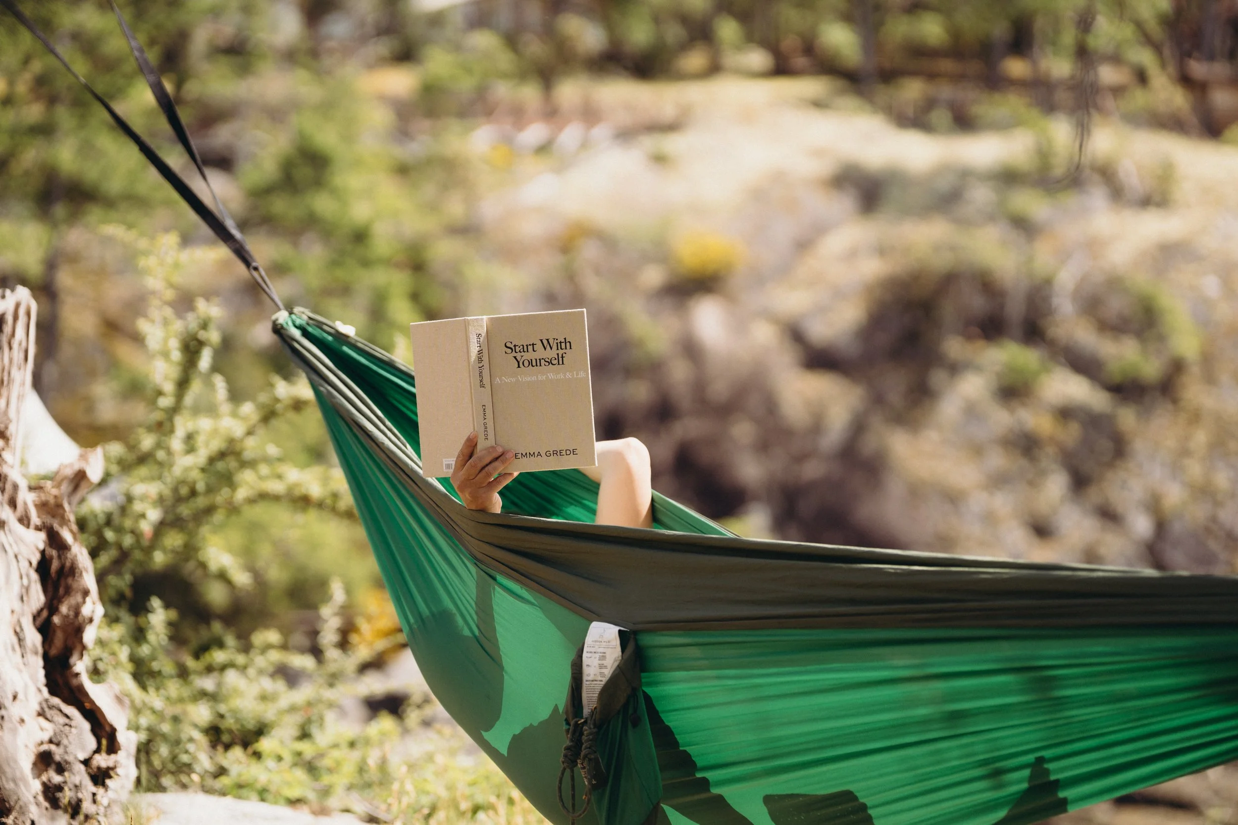 Person lying in a green hammock in a forest, reading a book titled 'Start With Yourself' by Emma Grede.