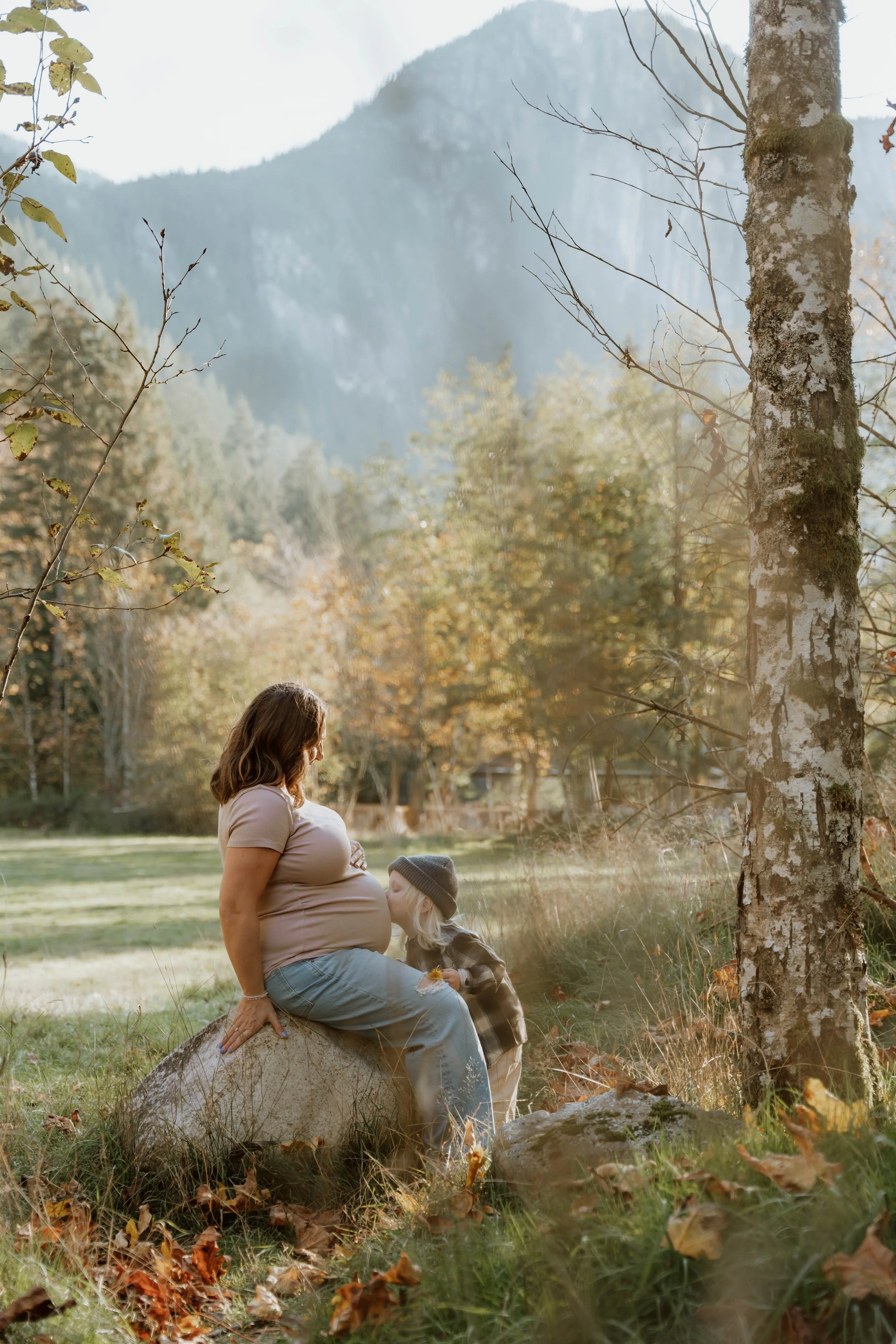 A pregnant woman sitting on a large rock in a forested area with a young girl, kissing her belly. The background shows trees and mountains, with sunlight filtering through the foliage, indicating autumn.