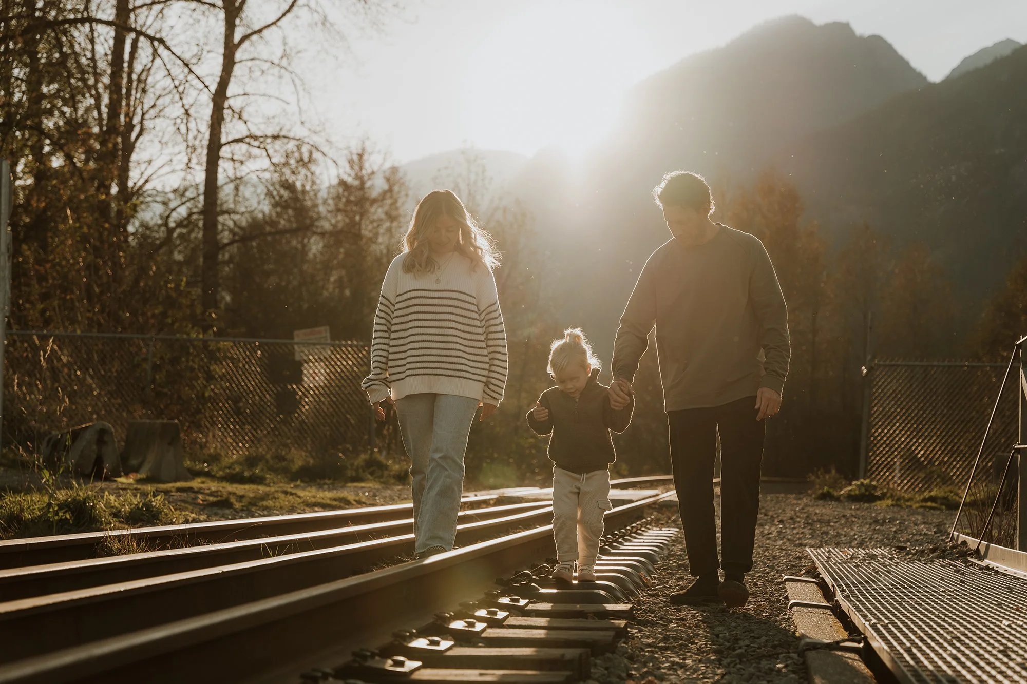 A family of three, with a young girl and her parents, walking hand in hand along a railway track during sunset in a natural, mountainous area.
