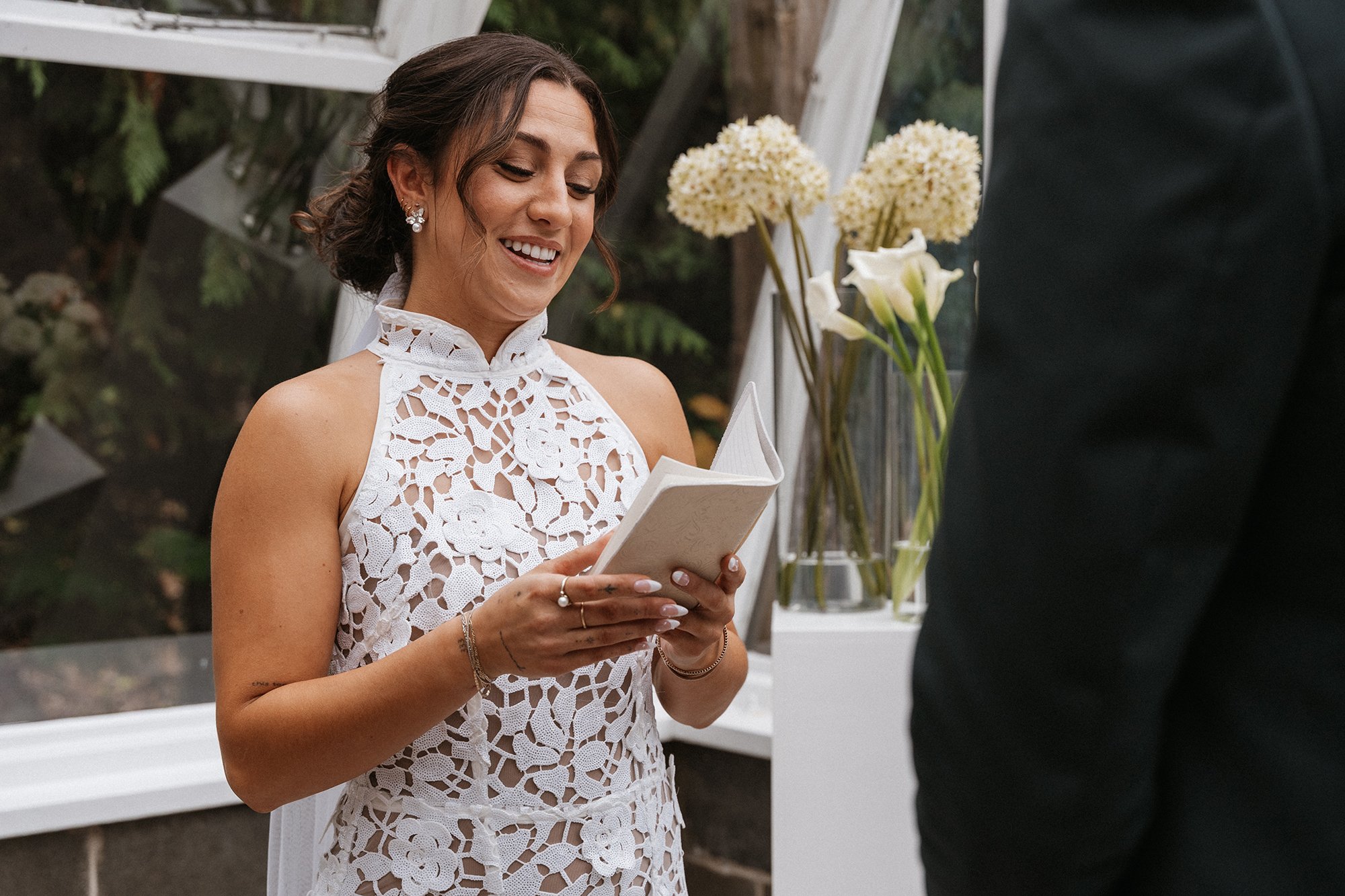 A woman in a white lace dress with jewelry, smiling, reading from a book during a wedding ceremony outdoors.