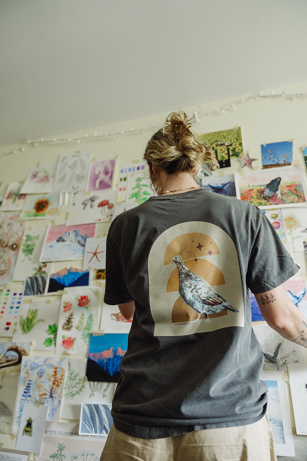 Person with curly hair tied in a bun, wearing a black T-shirt with a bird and sunset graphic, standing in front of a wall decorated with various colorful artwork and nature illustrations.