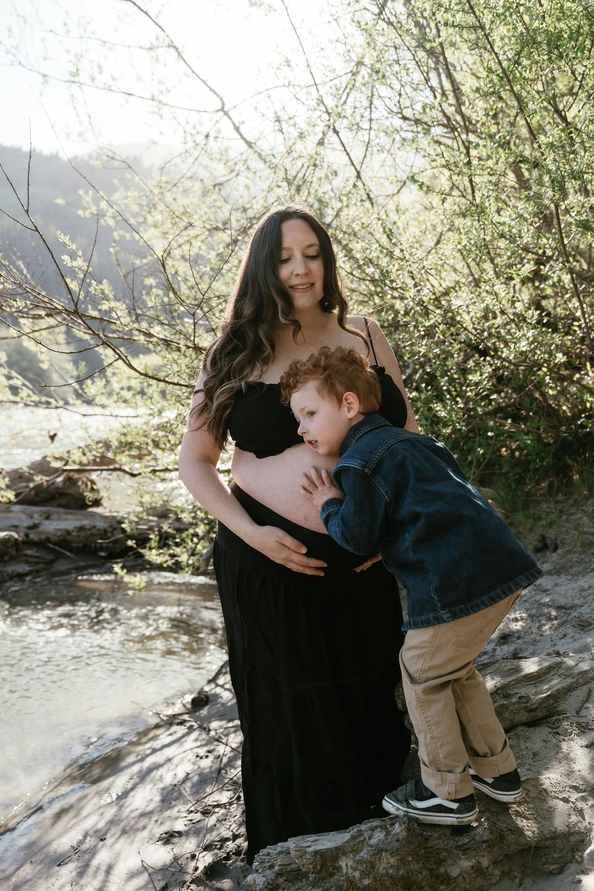 A woman with long dark hair and a young boy with curly red hair, both outdoors by a stream with leafless and green trees in the background, standing on a driftwood log.