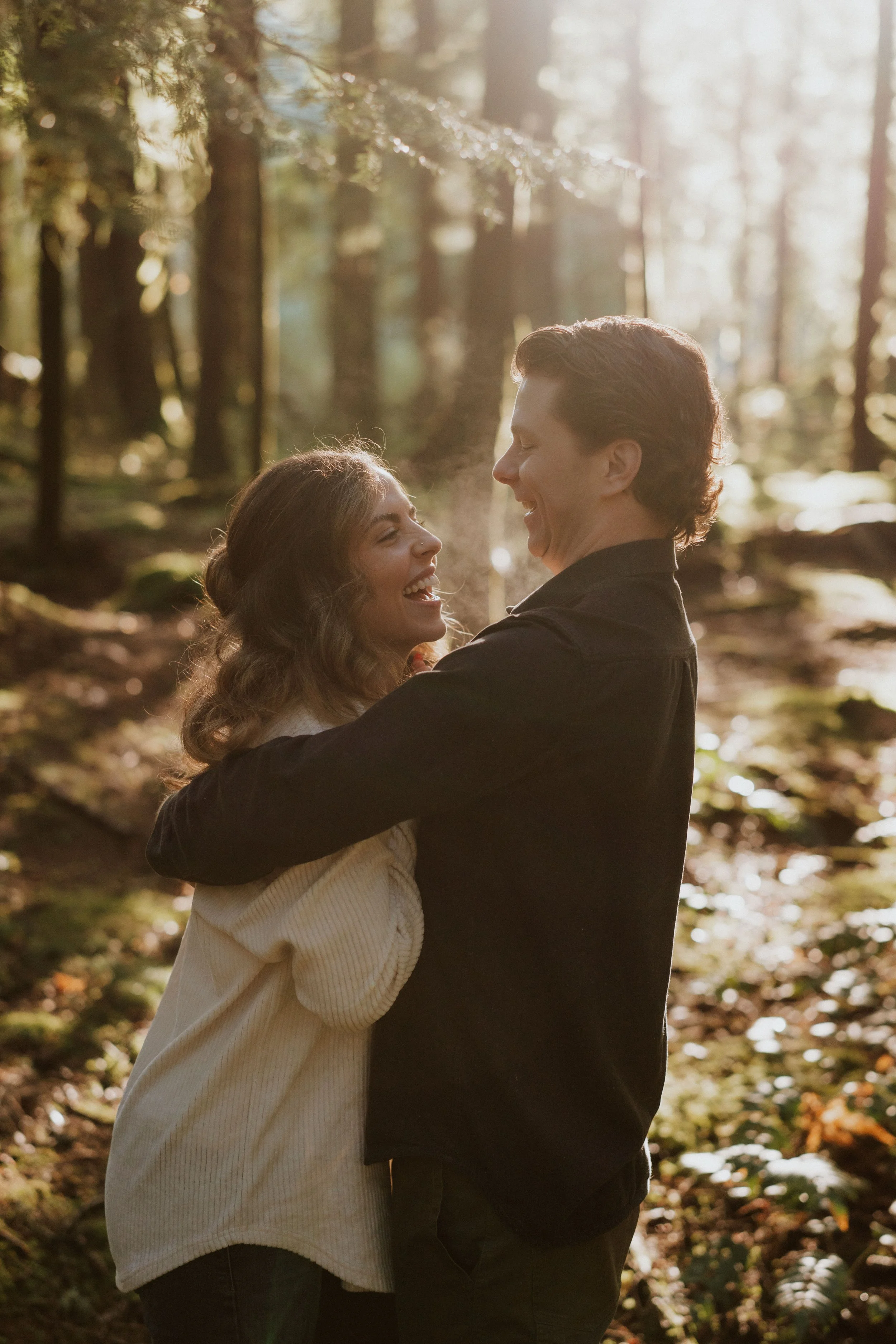 A couple happily embracing in a sunlit forest, with trees and fallen leaves in the background.