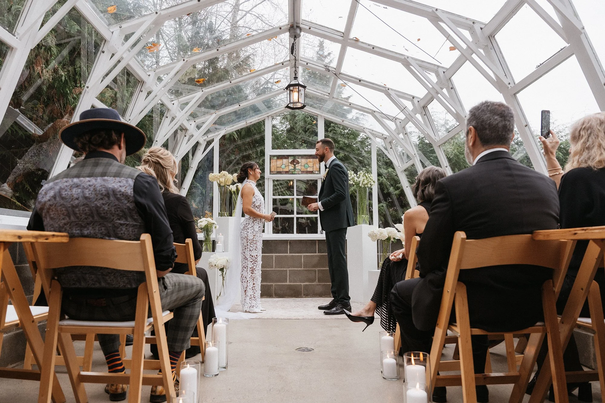 Couple exchanging vows at a wedding ceremony inside a glass greenhouse with wedding guests seated on wooden chairs.