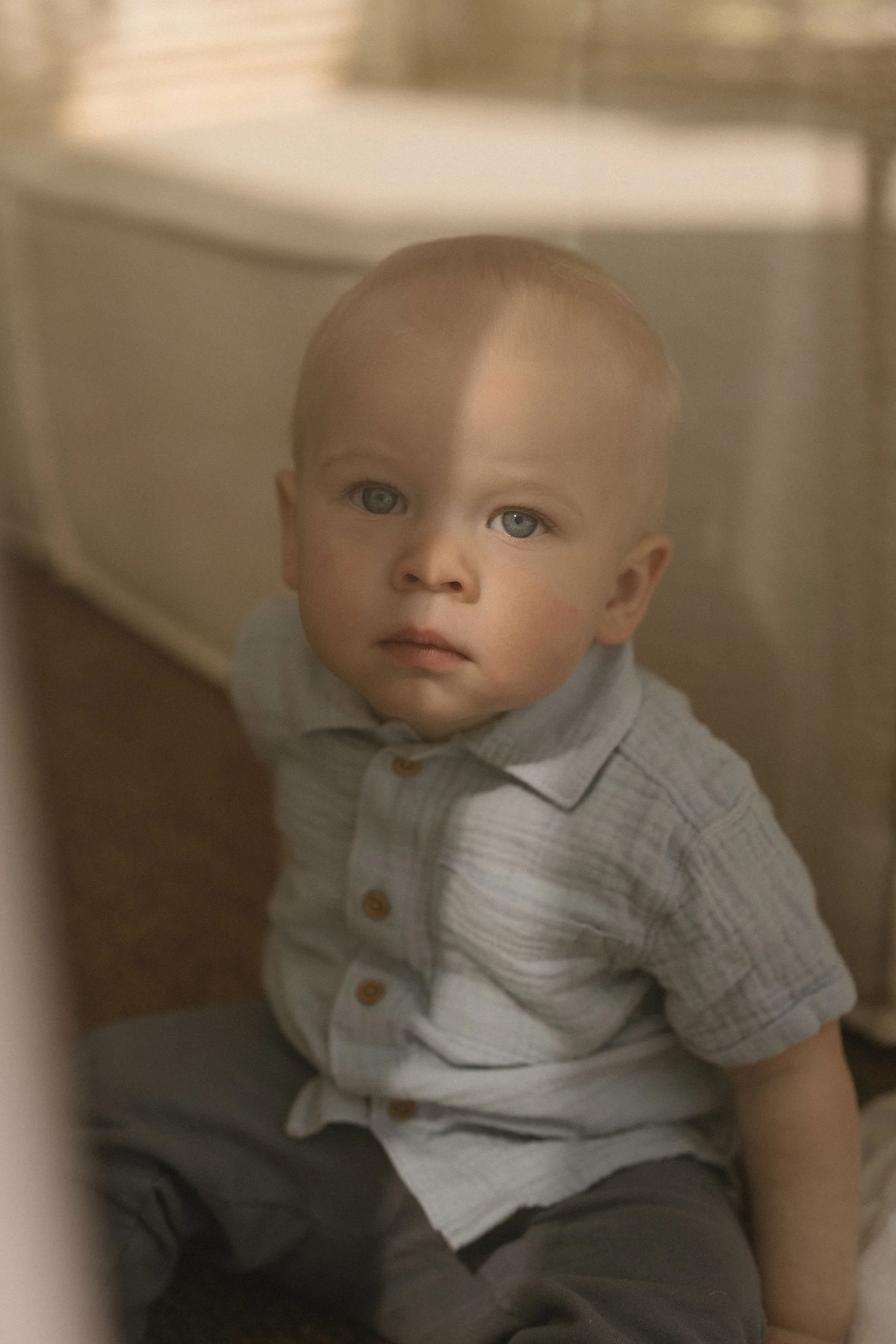 A young boy standing in a small, enclosed space, looking directly at the camera with a neutral expression. He has short, light brown hair and blue eyes, and is wearing a gray button-up shirt with rolled sleeves and dark pants.