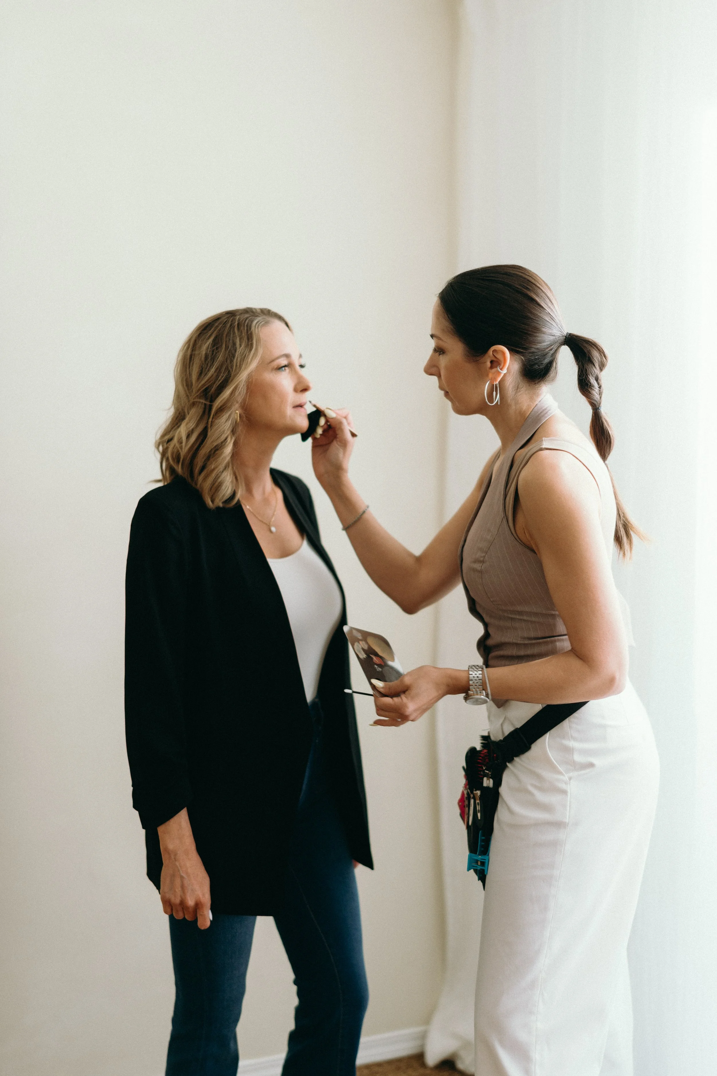 A makeup artist applying makeup to a woman standing against a plain light-colored wall.