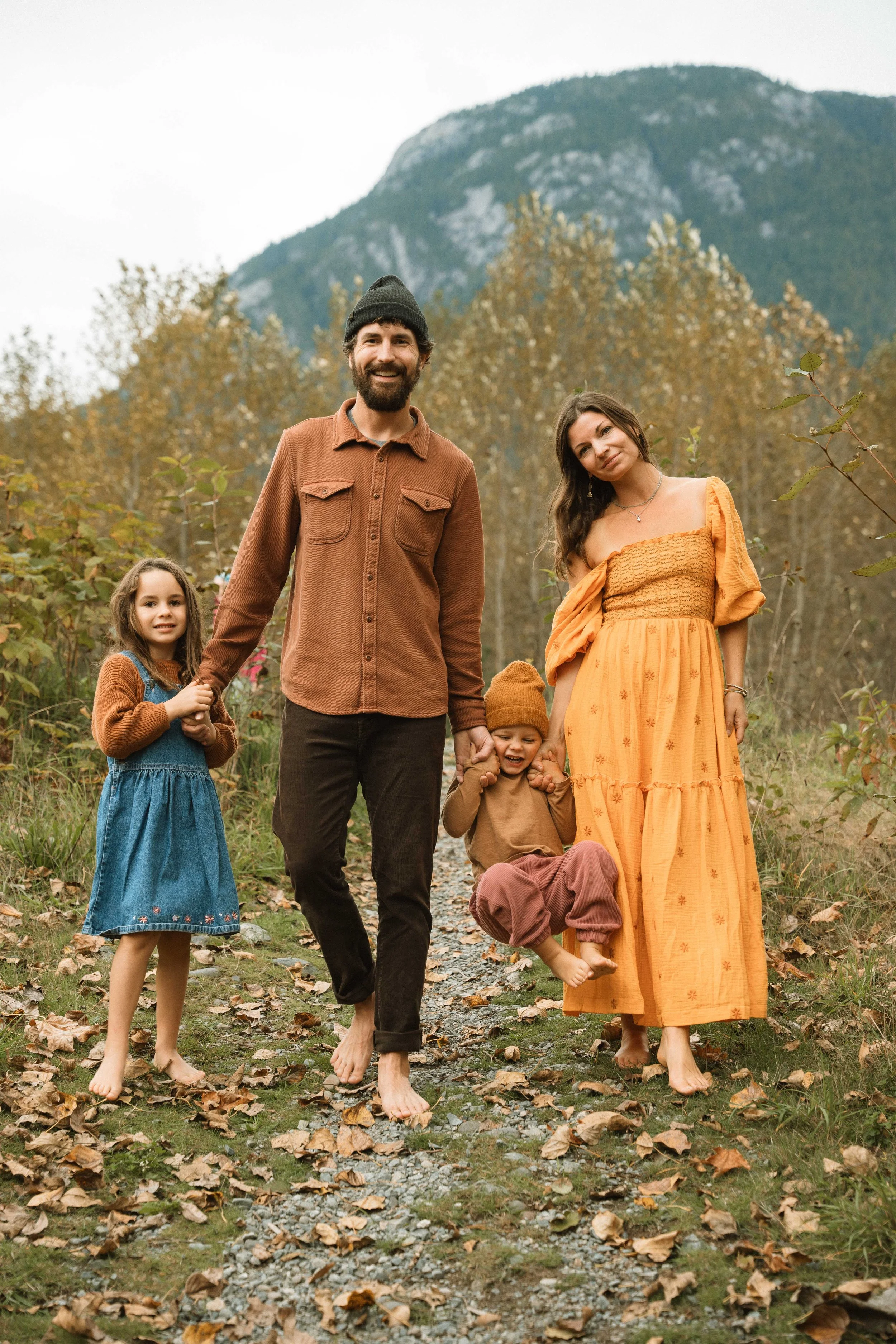 A happy family of four walking barefoot on a trail in a forested area during autumn with mountains in the background, holding hands and smiling.