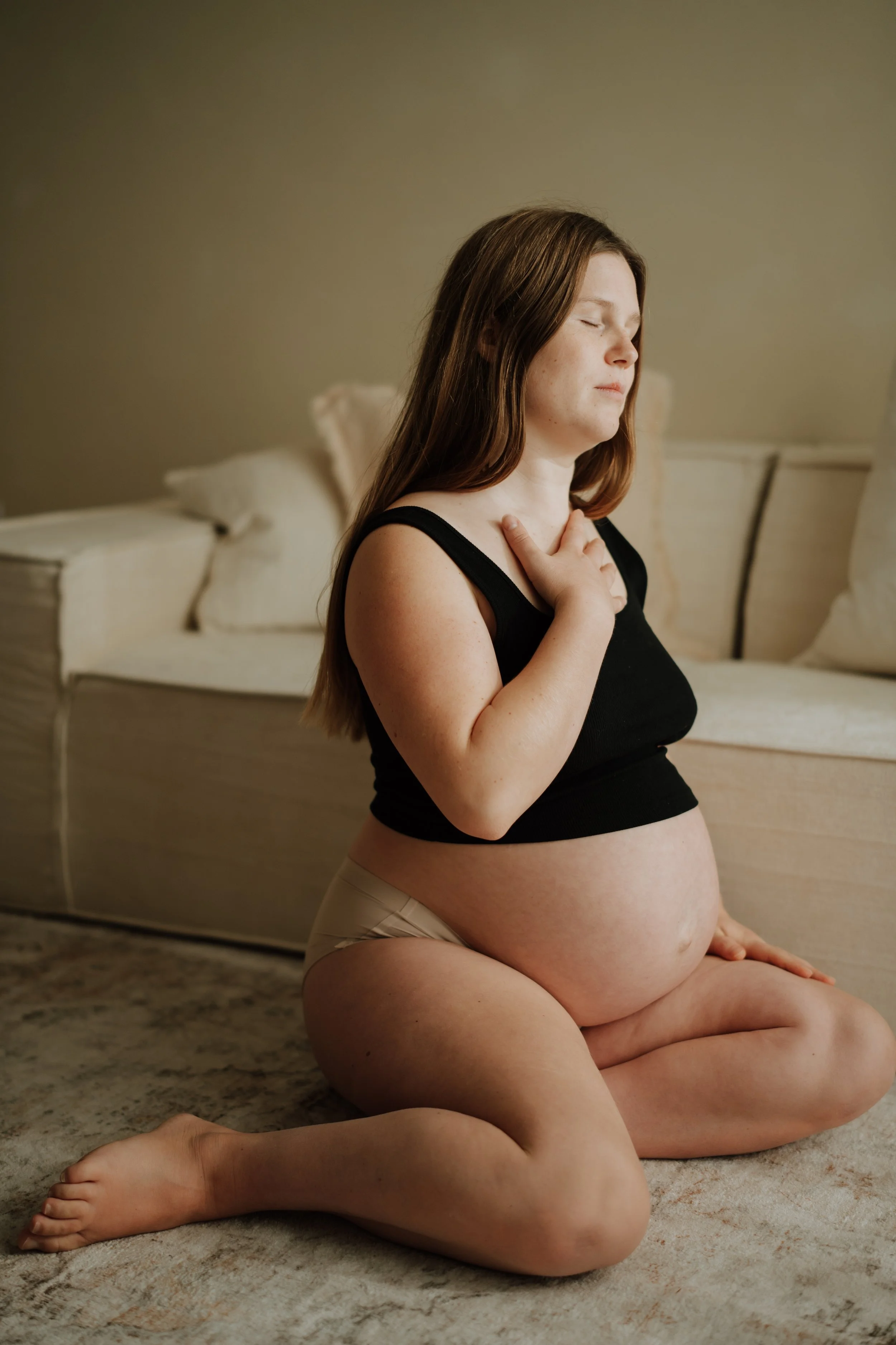 A pregnant woman with long brown hair sitting on a carpeted floor in a room with beige walls, eyes closed, hand on her chest, wearing a black sleeveless top and beige pants.