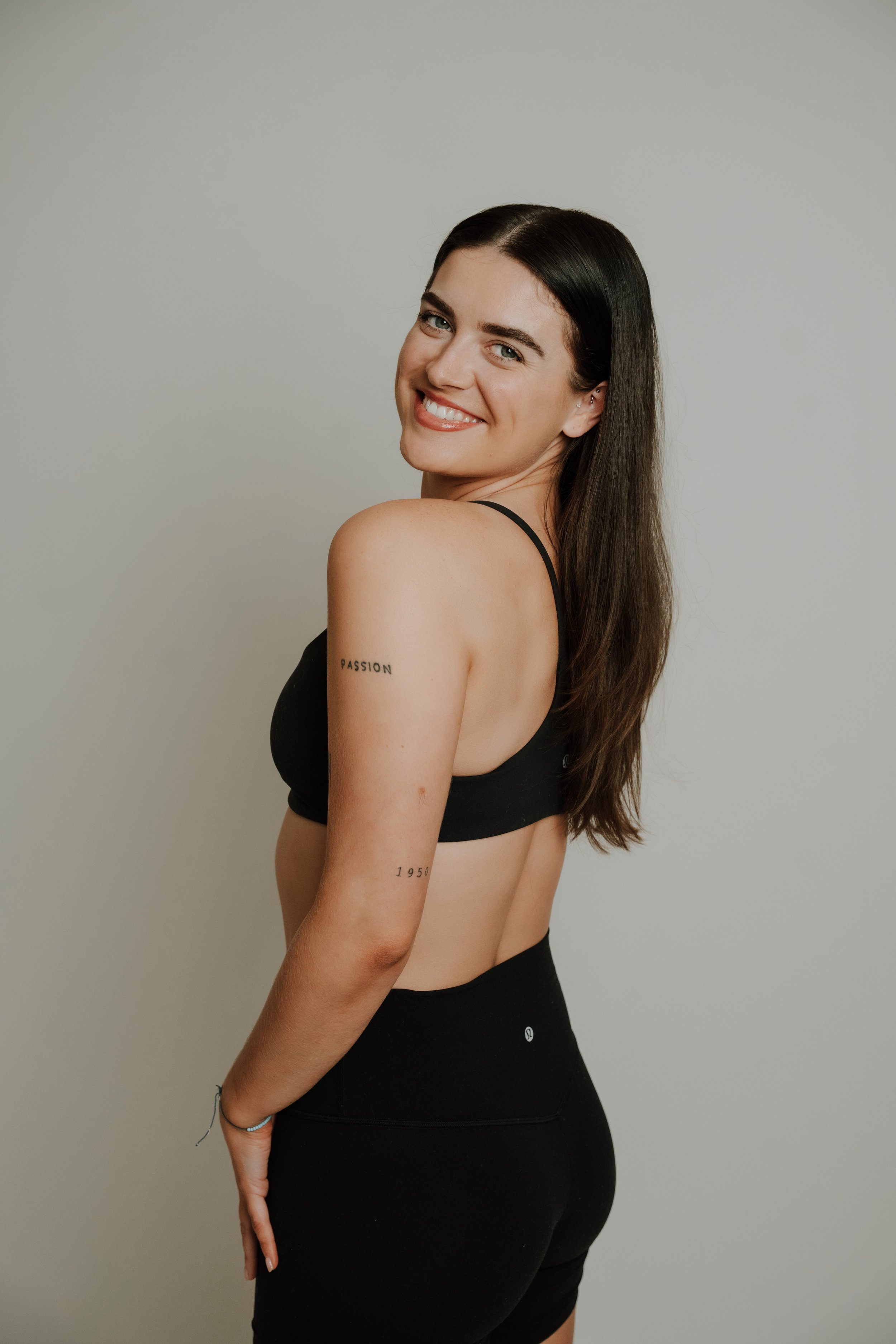 Smiling woman with long dark hair, wearing a black sports bra and black leggings, standing against a plain light-gray background, with tattoos on her left shoulder and arm.