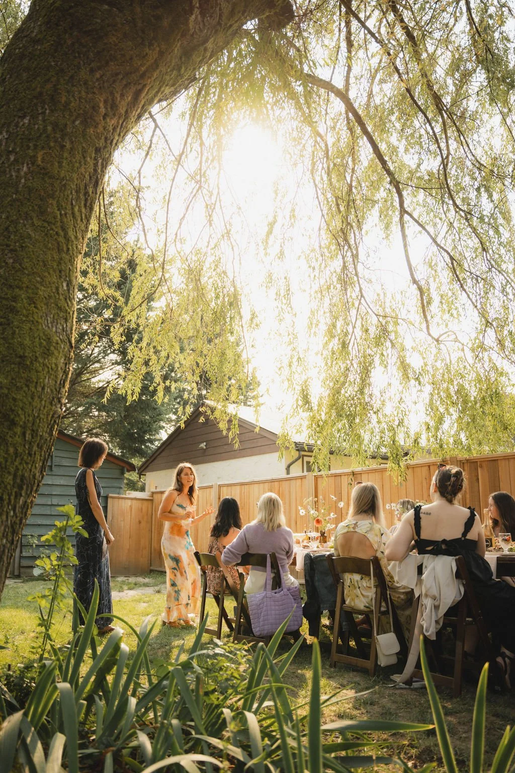 People gathered outdoors for a garden party or brunch, seated around a table with flowers and food, under a large tree with sunlight filtering through the leaves.