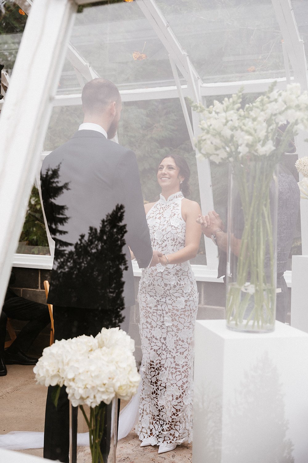 A bride and groom exchanging vows inside a glass greenhouse with white flowers.