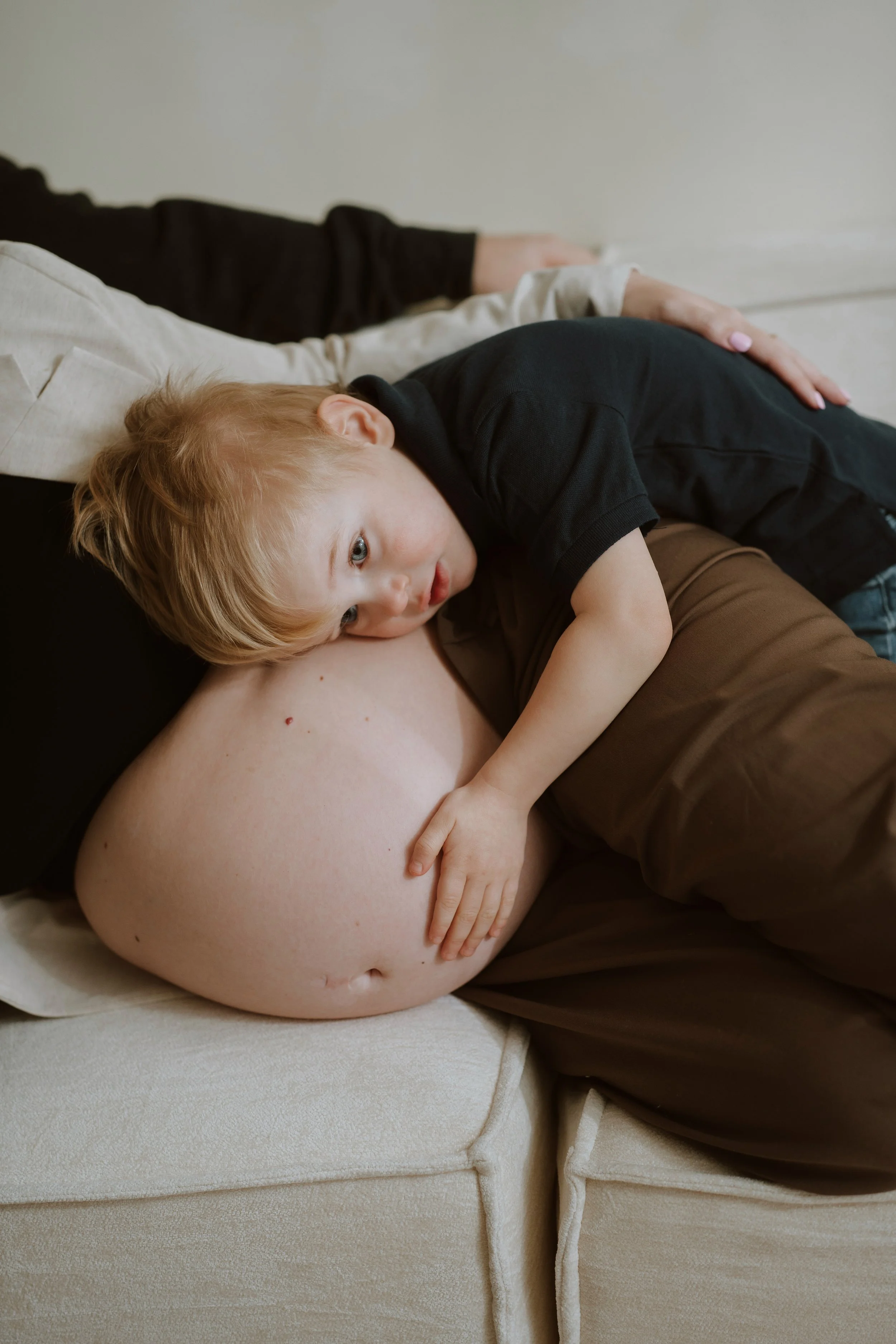 A young child with blonde hair and blue eyes lying on an adult's lap, holding onto their leg, with a large bald spot on the adult's head, sitting on a beige sofa.