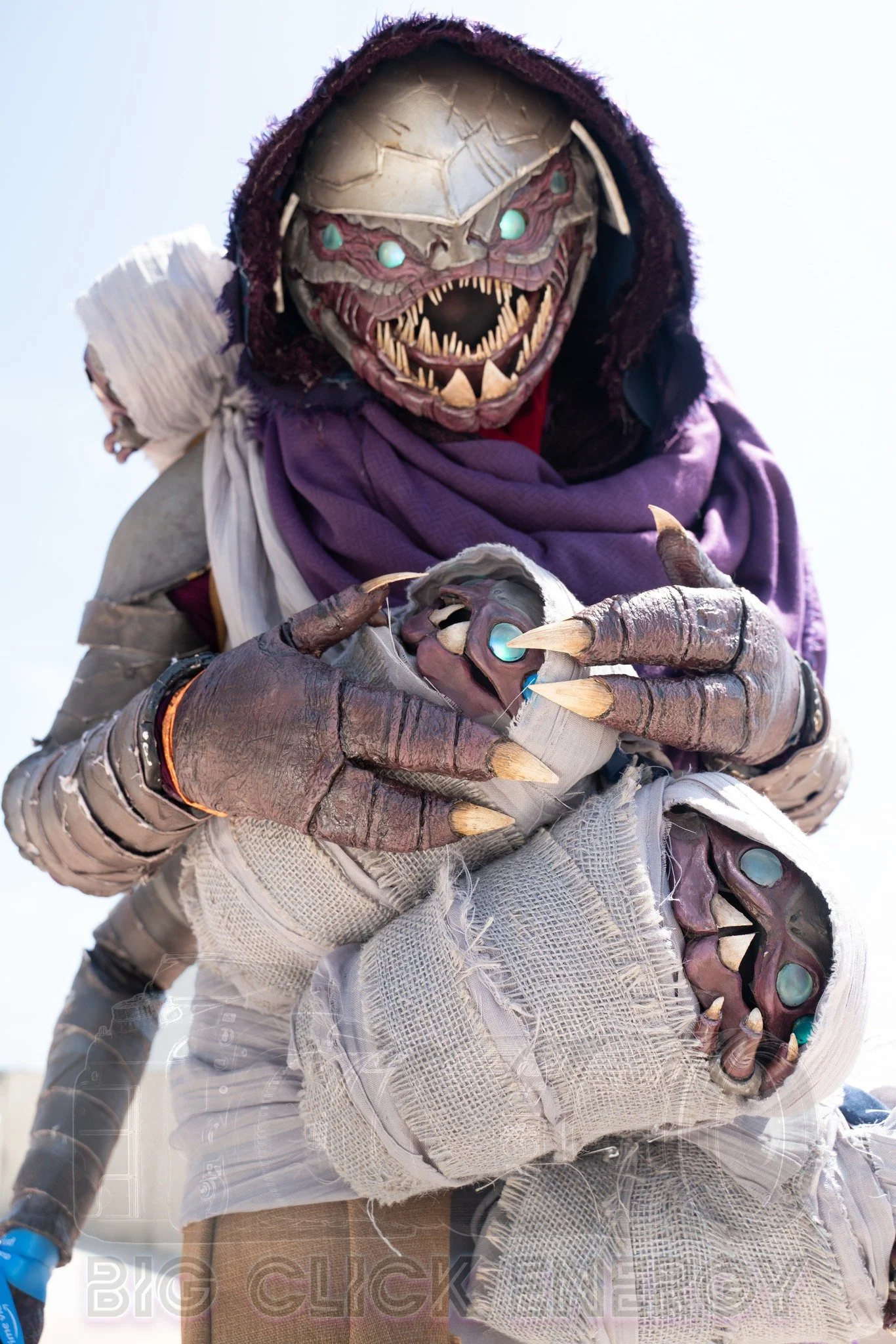 Cosplay of a Buglike alien in a purple hood with 4 glowing blue eyes, 2 claws on each hand, and 2 rows of needle-like teeth. It has 4 swaddled babies of the same species in carried in a baby sling