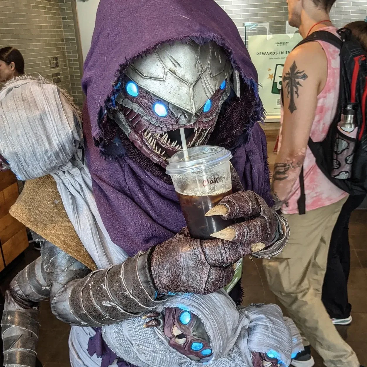 Cosplay of a Buglike alien in a purple hood with 4 glowing blue eyes, 2 claws on each hand, and 2 rows of needle-like teeth. It has 4 swaddled babies of the same species in carried in a baby sling, and holds an iced coffee with a straw up to its open