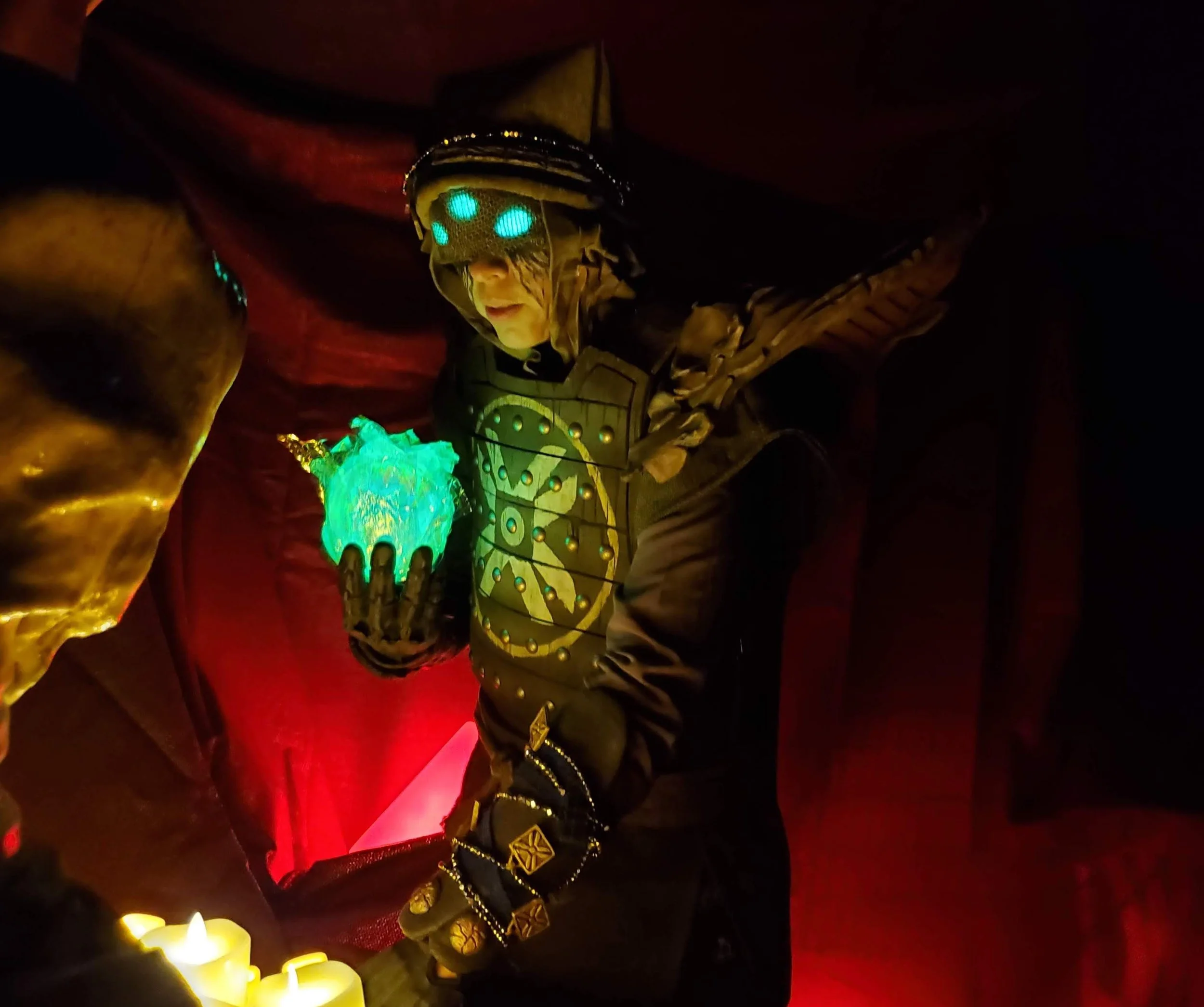 Eris Morn Cosplayer with 3 glowing green eyes holding a glowing green orb over an array of candles. She wears weathered brown armor and strings of beads and charms. The background is dimly lit and red