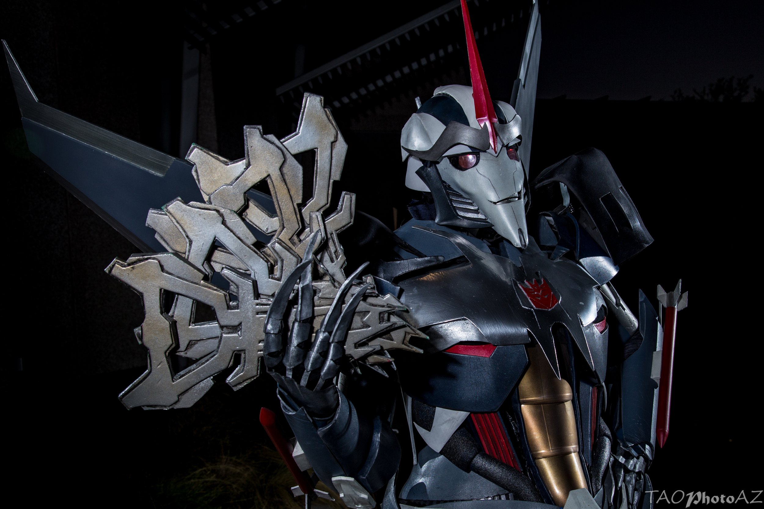 Cosplay of Transformers Prime Starscream, a slender robot with a pointed face and long jet wings. He has red details and eyes, a smug expression, and holds 4 key-like artifacts in his talons. Both forearms have missiles attached