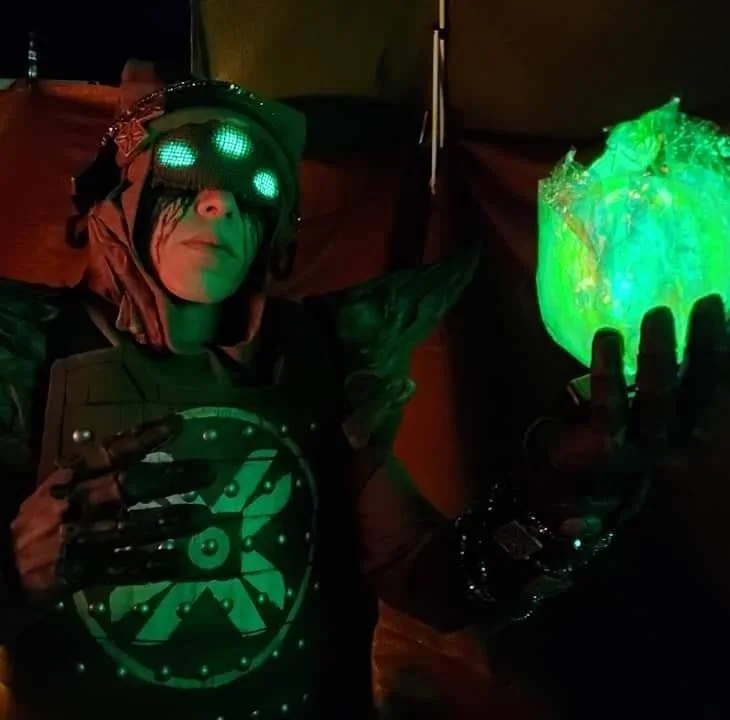 Eris Morn Cosplayer with 3 glowing green eyes holding up a glowing green orb. Black tears flow from under a veil and she wears weathered brown armor. The background is dimly lit and red