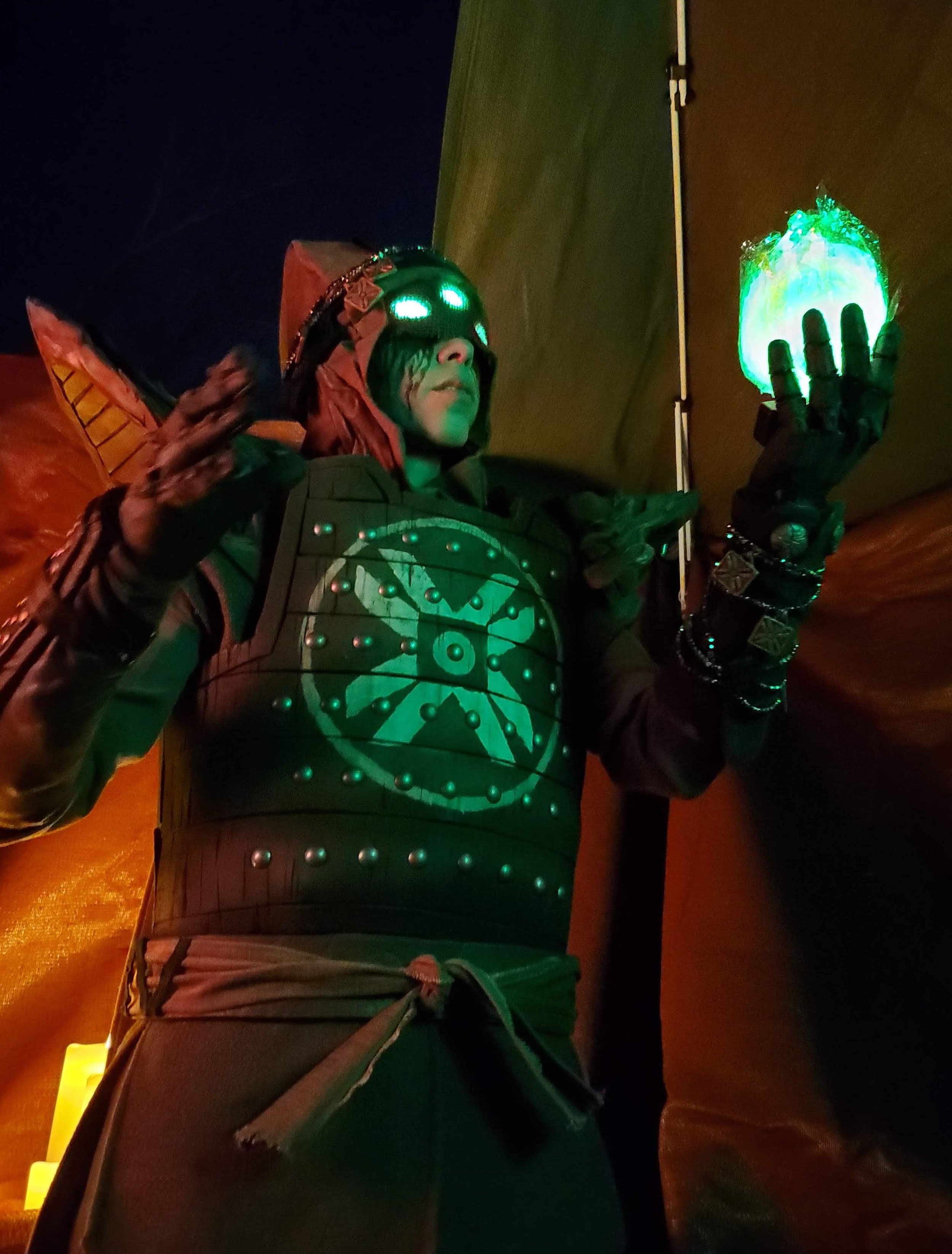 Eris Morn Cosplayer with 3 glowing green eyes holding up a glowing green orb . She wears weathered brown armor and strings of beads and charms. The background is dimly lit and red