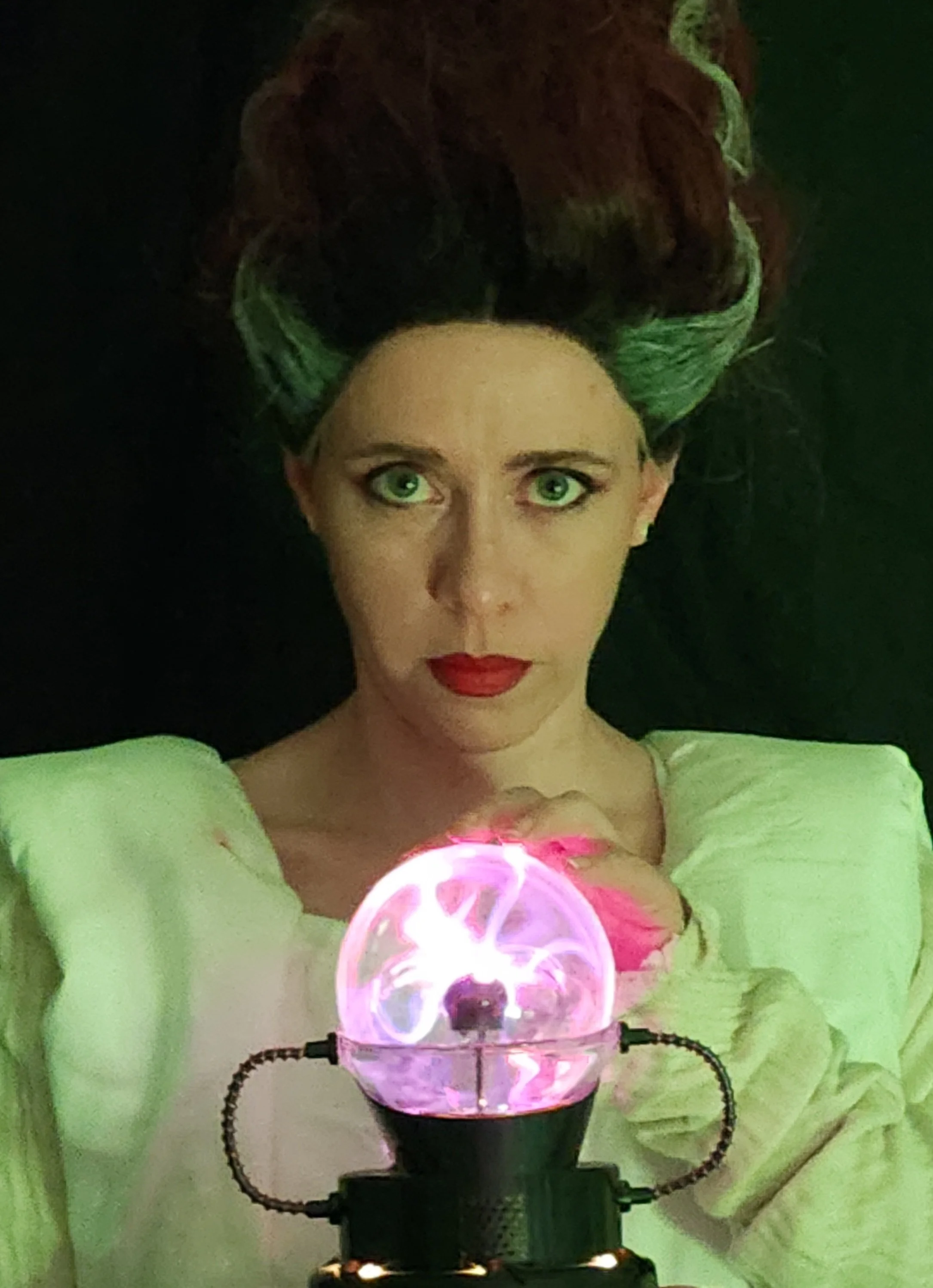 Cosplay of the Bride of Frankenstein in a white sheet gown and a dark red beehive wig with white streaks on the sides. She stares straight at the viewer and holds a plasma sphere with pink electric arcs inside