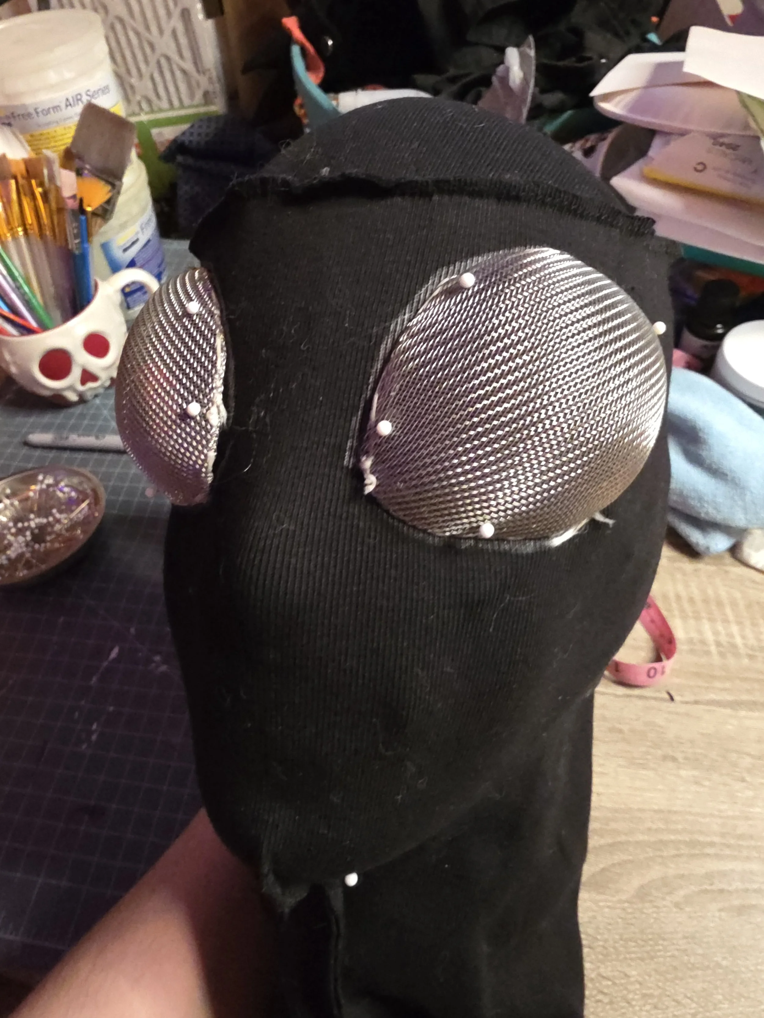 Metal mesh egg-shaped eye covers pinned in place on the front of a black fabric ninja's hood. The hood is inside out and the eye outline is traced in sharpie.