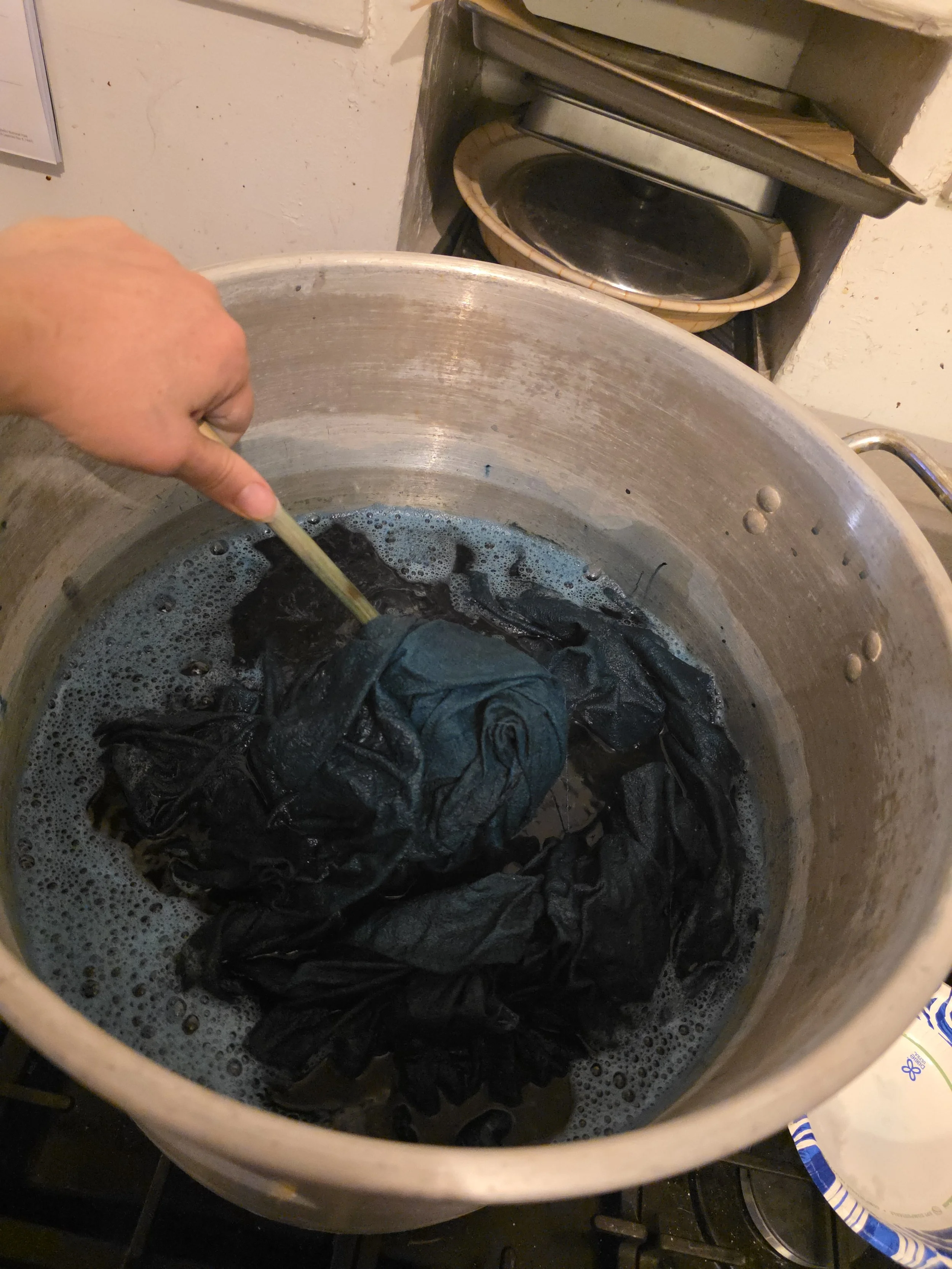 53 quart tamale pot sitting on 3 of the 5 burners on a gas stove. It is more than half full of dark green dye, and a hand stirs fabric boiling in it with a wooden spoon 