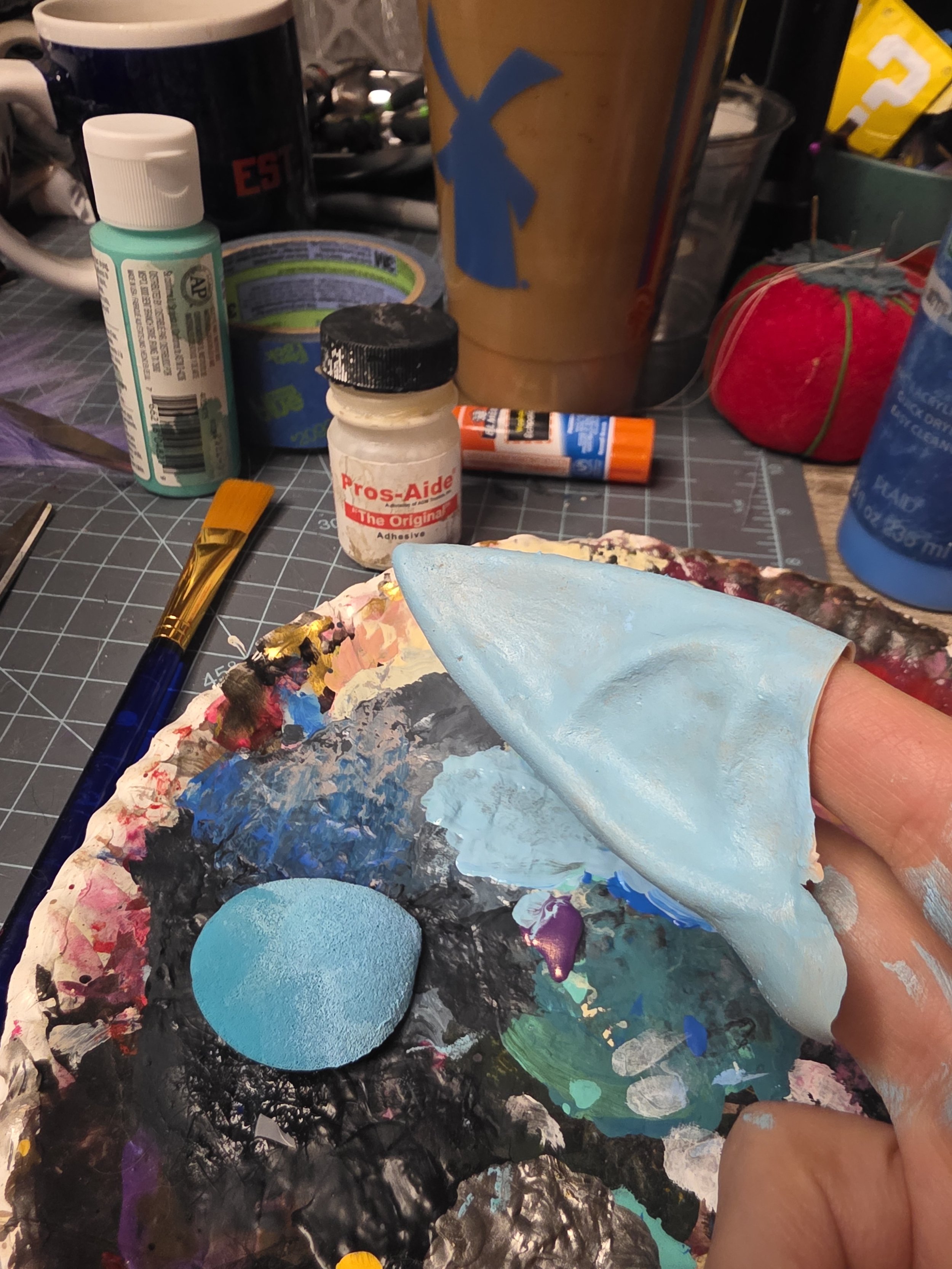 latex pointy ear held on 2 fingers, painted pale blue. paint is mixed on a paper plate, with a bottle of Pros-Aide, because that is how paint sticks to latex