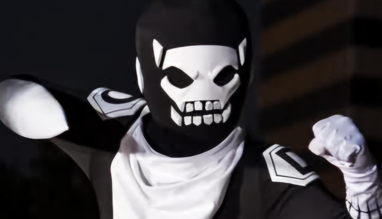 Cosplay of a tokusatsu footsoldier in a black bodysuit with a white neck kerchief and shoulder badges with D on them. The head is round with a skull-like face. One hand has a white glove and the other is inside a sword-like appendage