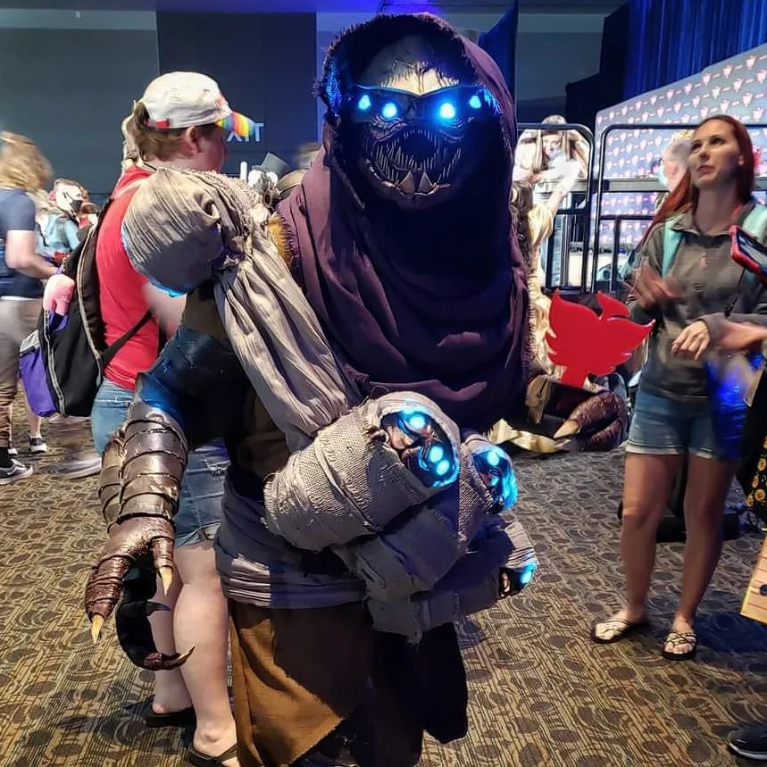Cosplay of a Buglike alien in a purple hood with 4 glowing blue eyes, 2 claws on each hand, and 2 rows of needle-like teeth. It has 4 swaddled babies of the same species in carried in a baby sling. One hand holds a red Phoenix-shaped cosplay contest 